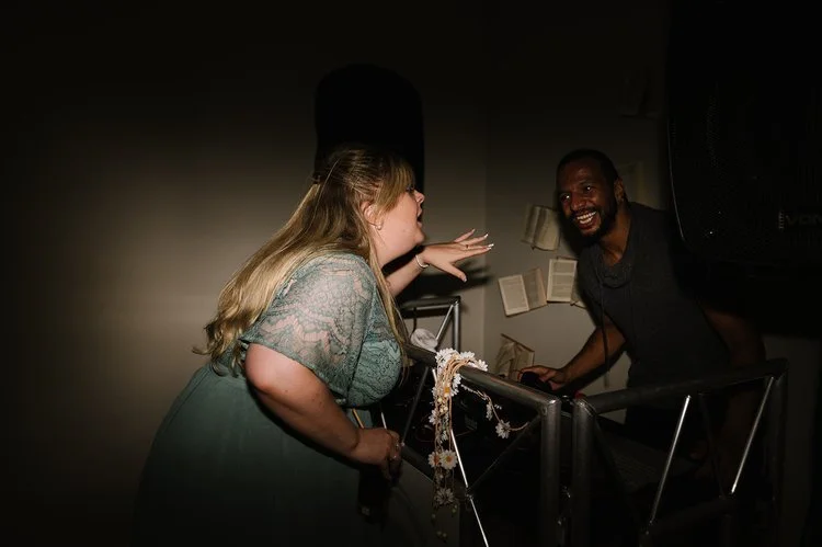 A wedding guest shares a fun moment with The Best Men DJ during a lively wedding reception. The DJ booth, decorated with flowers and creative open-book décor, sets the scene for an unforgettable evening of music and laughter.