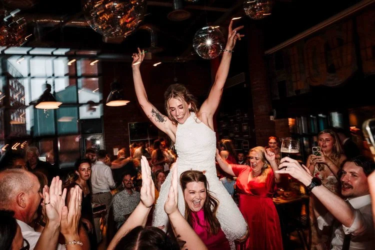 A vibrant and joy filled wedding moment captured by Amy Faith Photography, showing the couple celebrating outside with a lively brass band. The bride holds a bold, colourful bouquet while laughing in the middle of the musicians, who are performing wi