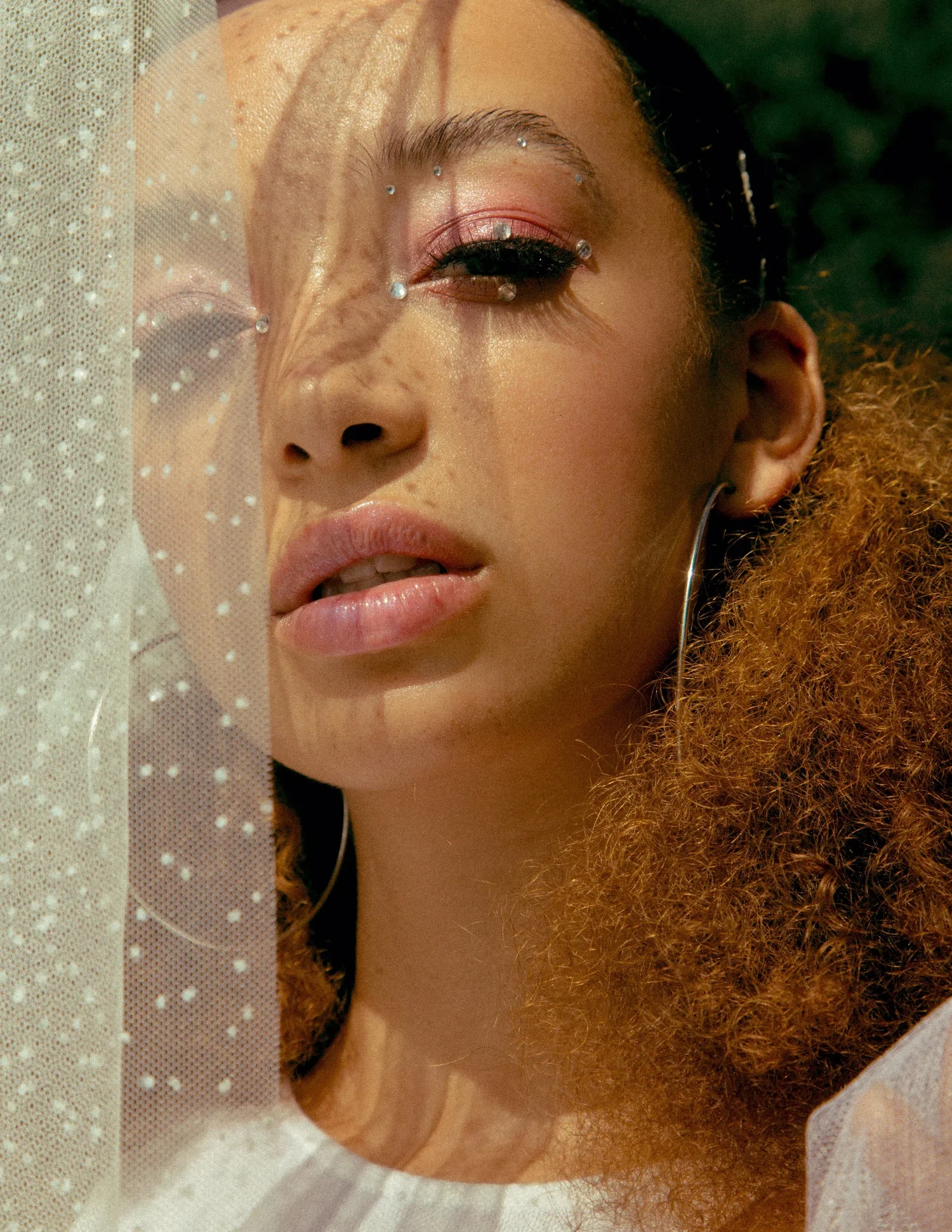 A creative and romantic bridal look by Powder Blue Artistry - featuring soft pink eyeshadow, glossy nude lips, and crystal embellishments delicately framing the eyes for a celestial touch. The sheer dotted veil adds texture and mystery, while the nat