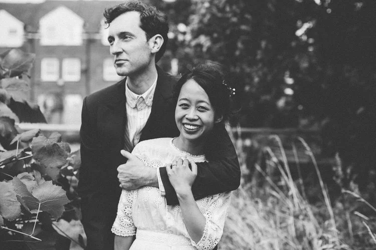 A joyful black-and-white wedding portrait captured by Mirrorbox Photography, showing a couple embracing in a relaxed outdoor setting surrounded by soft foliage and long grasses. The bride’s vintage-inspired lace dress and radiant smile bring warmth a
