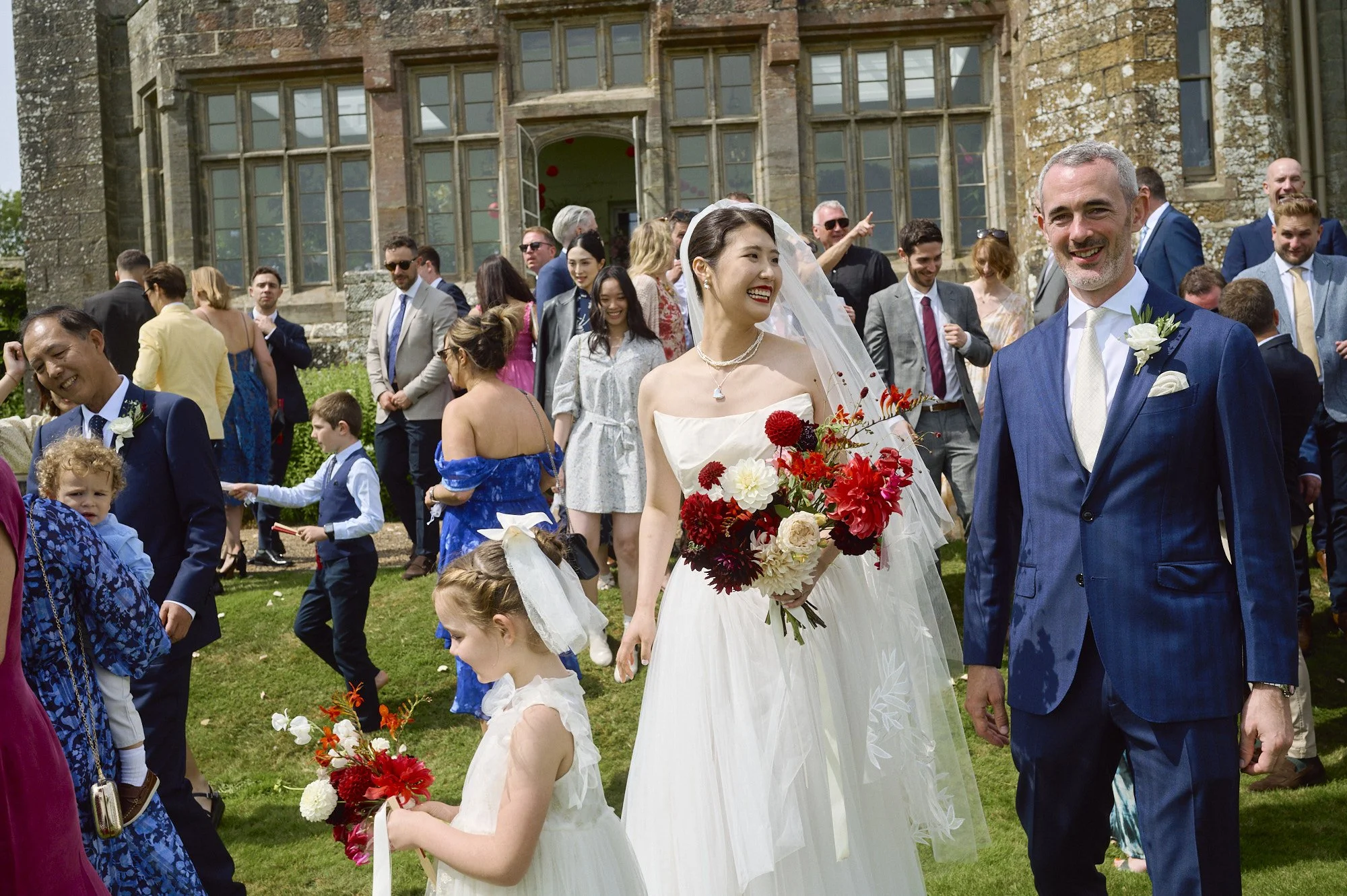 A lively and cinematic wedding moment captured by Wayne La Photography, showing the couple sharing a kiss while their guests react with joy and surprise. The groom’s navy suit and the bride’s voluminous white gown fill the foreground, creating an int