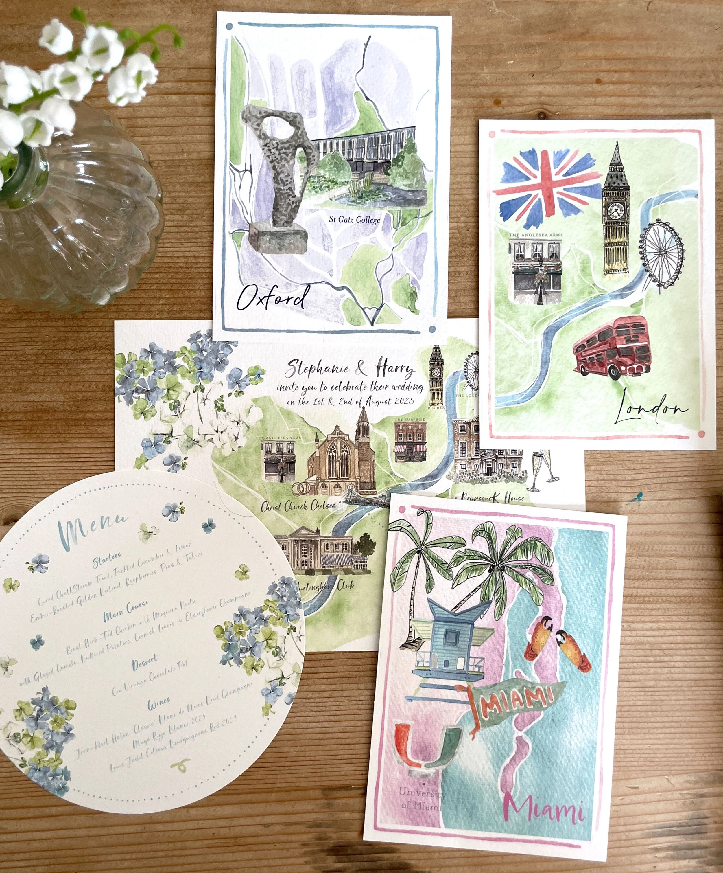 A vibrant collection of bespoke illustrated wedding stationery by Feather & Ink Stationery featuring personalised location artworks including Oxford, London and Miami alongside a floral circular menu and a detailed venue map all arranged on a rustic 