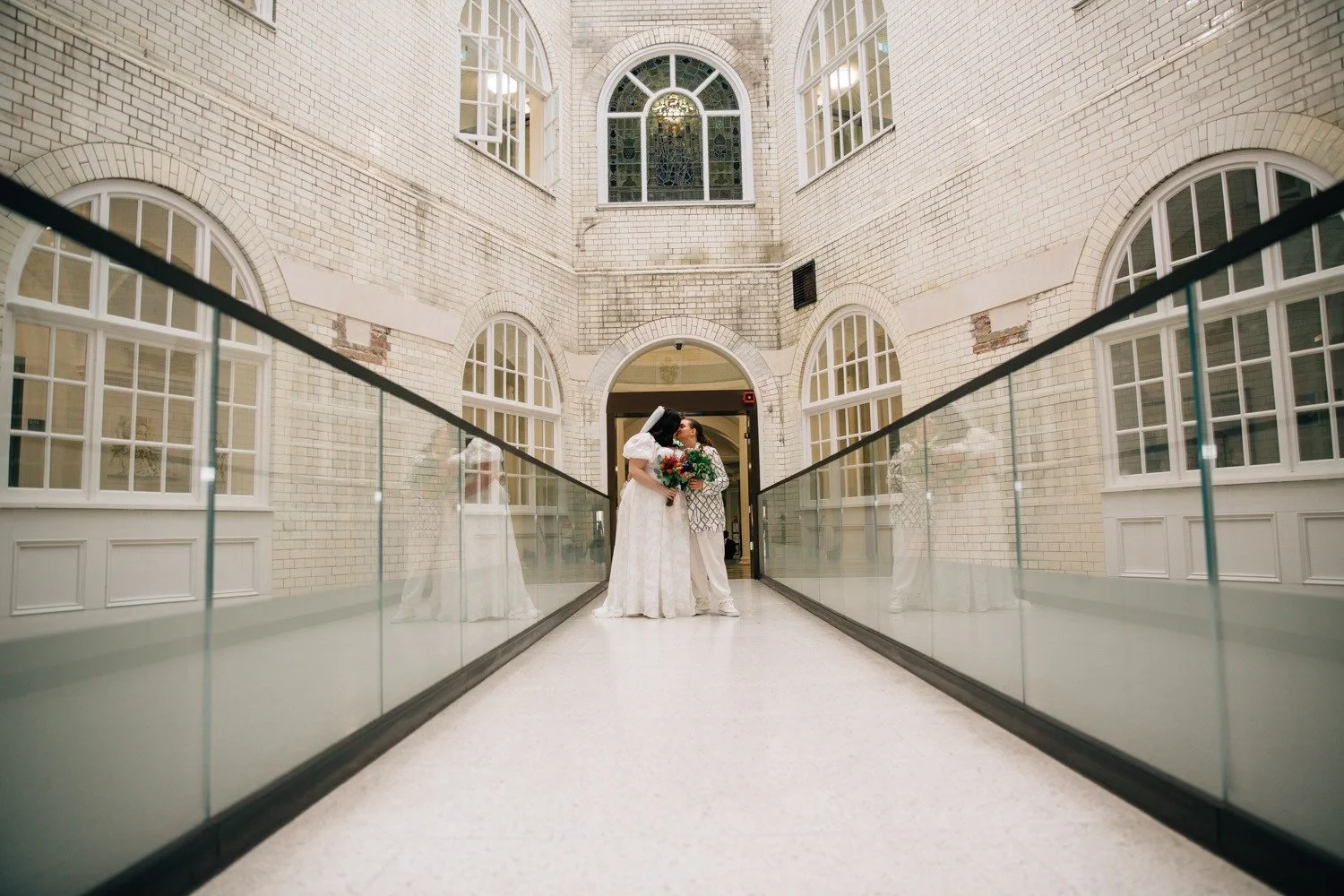 A romantic wedding portrait captured by Paola de Paola Photography, featuring two brides standing together on a modern glass walkway inside a historic white-brick building. One bride wears a flowing lace gown and veil, while the other wears a pattern