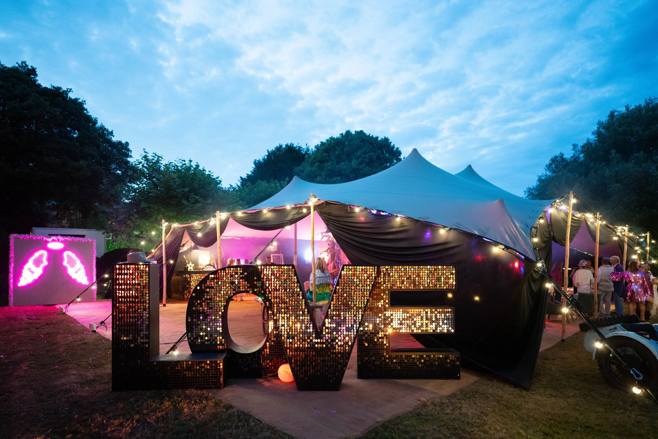 Outdoor event setup featuring a large illuminated ‘LOVE’ sign with shimmering sequins and warm lights in front of a stylish stretch tent. The evening scene is decorated with fairy lights, glowing pink neon angel wings, and guests enjoying a festive c