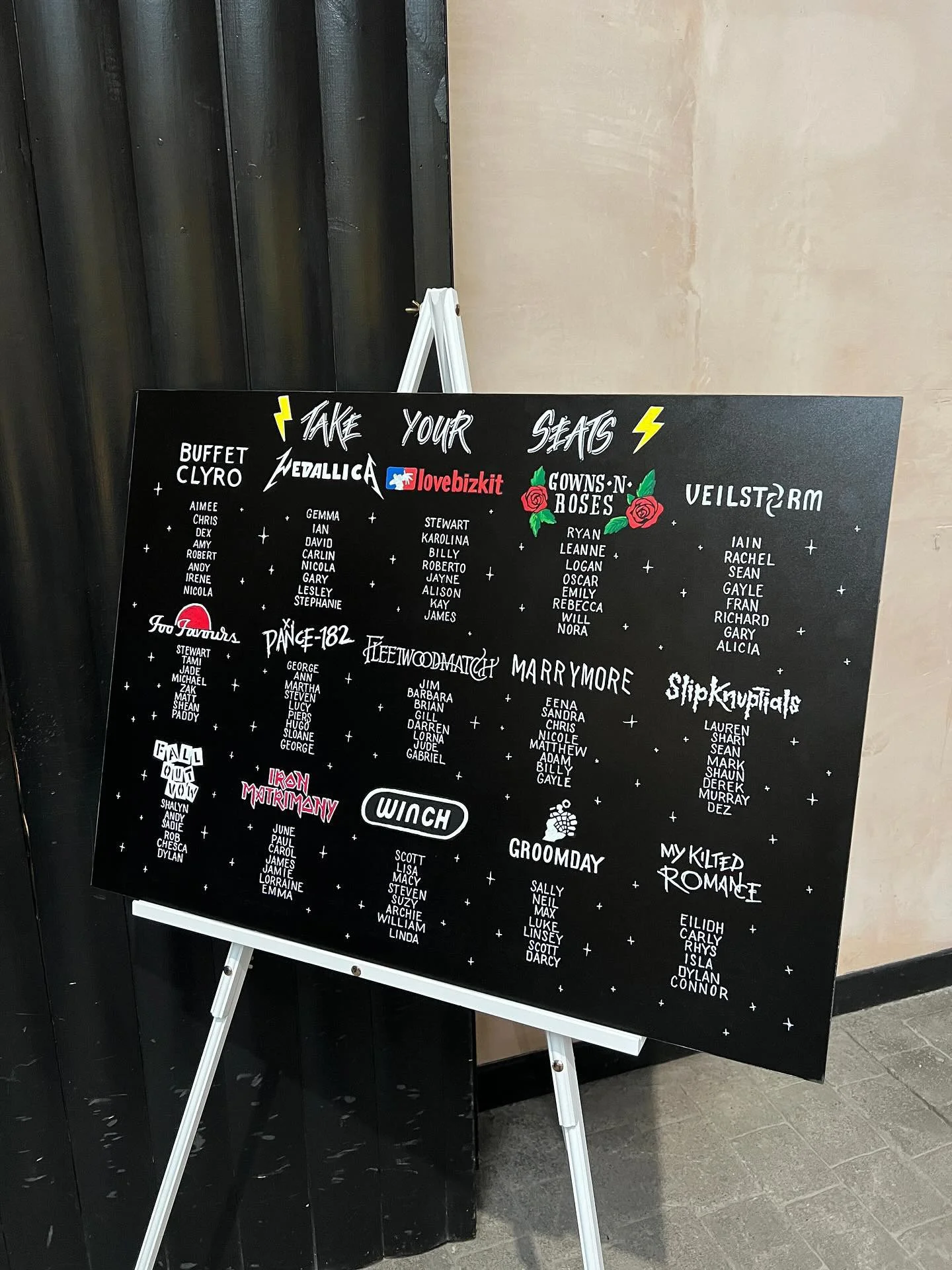 A bold, hand-illustrated wedding seating plan featuring a black background with playful, rock-inspired typography, custom icons and themed table names. Designed and painted by Toasty Type, the sign combines bespoke lettering, illustrated details and 
