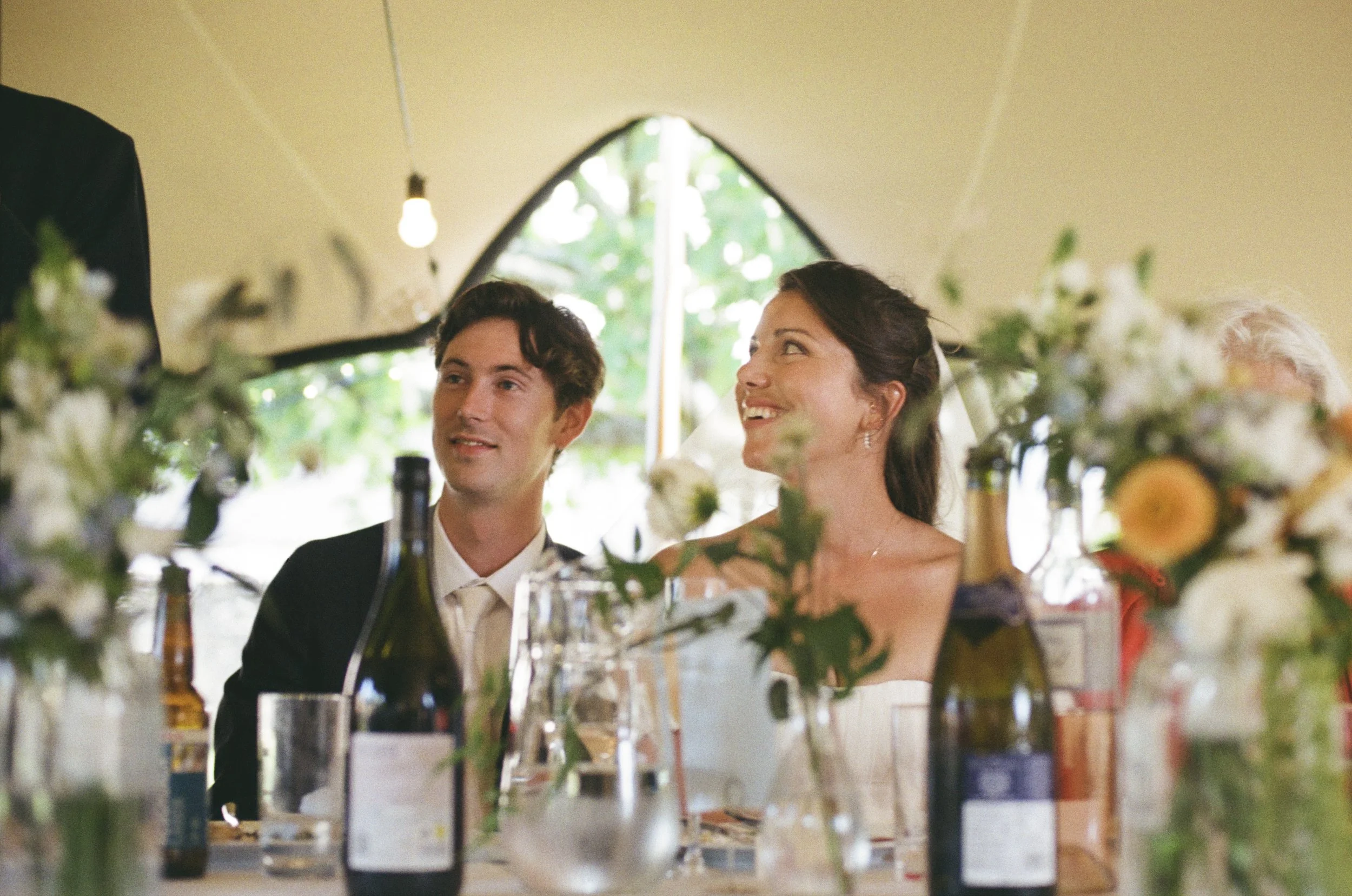 A couple sit together during wedding speeches inside a beautifully decorated marquee, captured by Roz Pike Photography. Surrounded by flowers, soft light and celebratory details, their joyful expressions create a warm candid moment that reflects the 