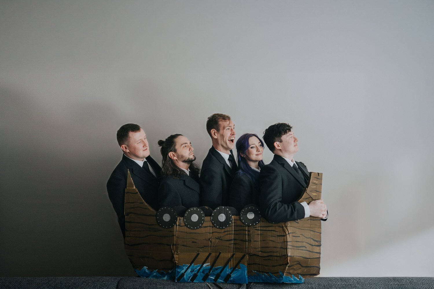 A playful, alternative wedding portrait showing the wedding party dressed in black suits, squeezed together inside a handmade cardboard boat prop, capturing the irreverent humour and creative storytelling style of Good Luck Wolf wedding photography i