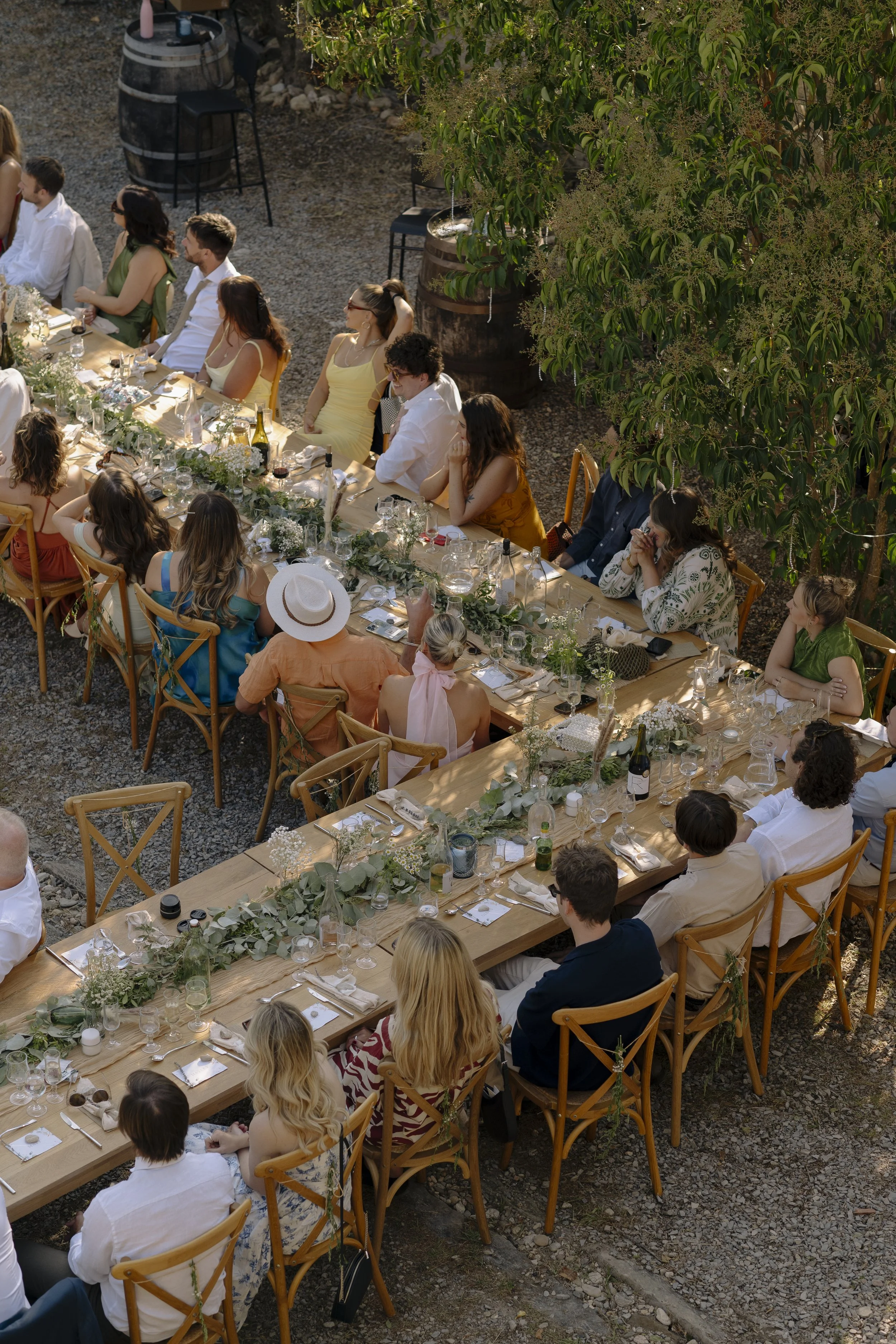 Guests gather around long wooden tables decorated with greenery and candles at an outdoor wedding feast, captured by Roz Pike Photography. The warm golden light, relaxed seating and rustic setting create a beautiful candid scene from a modern alfresc