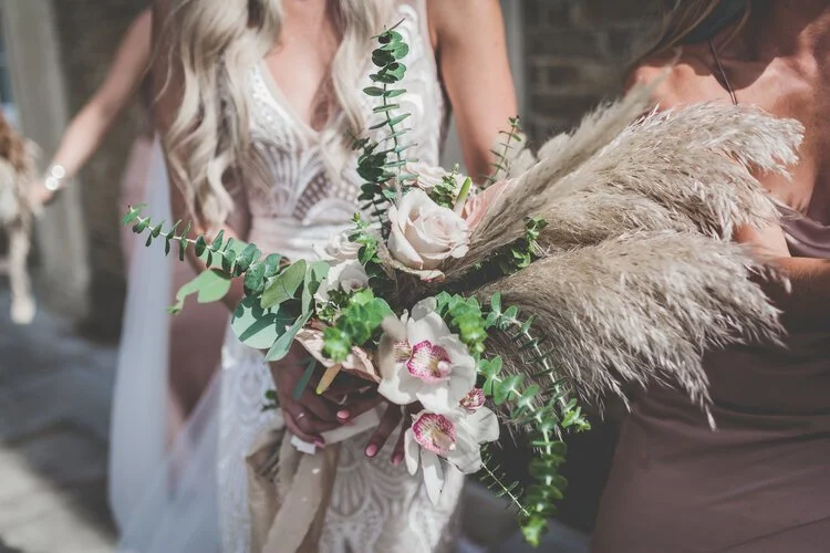 Modern boho wedding bouquet by Catherine Short Florals featuring soft blush roses, pampas grass, orchids and fresh eucalyptus. Held against an intricate lace gown in natural sunlight, this textured airy design highlights Catherine Short Florals’ cont