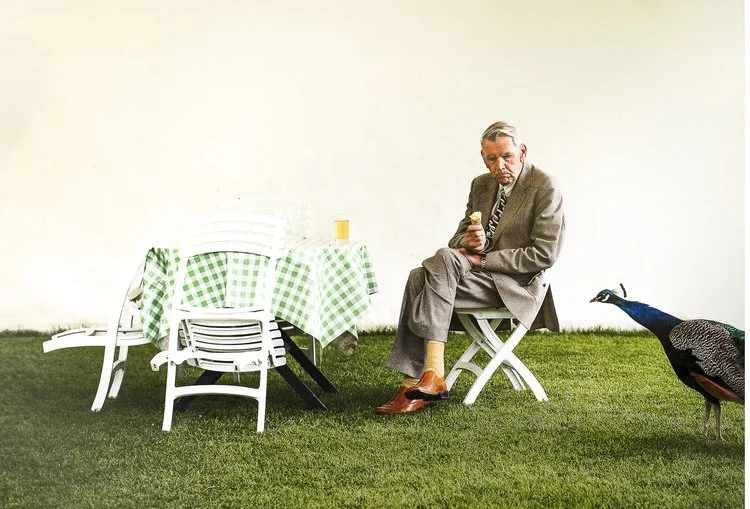 A quirky, cinematic scene with the offbeat humor typical of Matt Parry’s storytelling style. A man in a light-colored suit sits alone on a small white folding chair in the middle of a bright green lawn. He holds an ice cream cone and looks toward a p