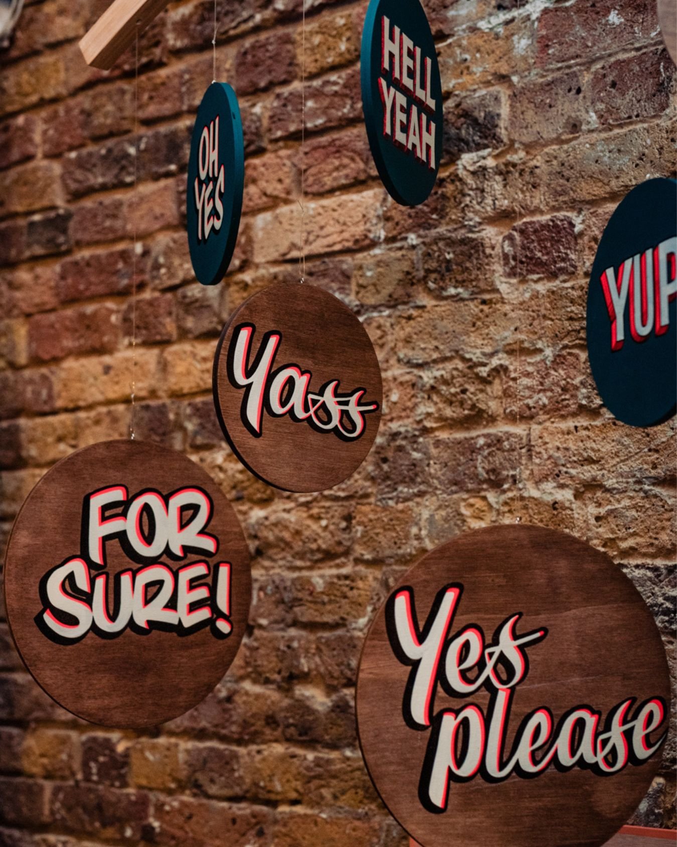 Close-up of bespoke Sylvie Signs wedding signage featuring circular wooden hanging signs with hand-painted lettering reading “Oh Yes,” “Hell Yeah,” “Yass,” “Yes Please” and “For Sure,” displayed against a rustic exposed brick wall.