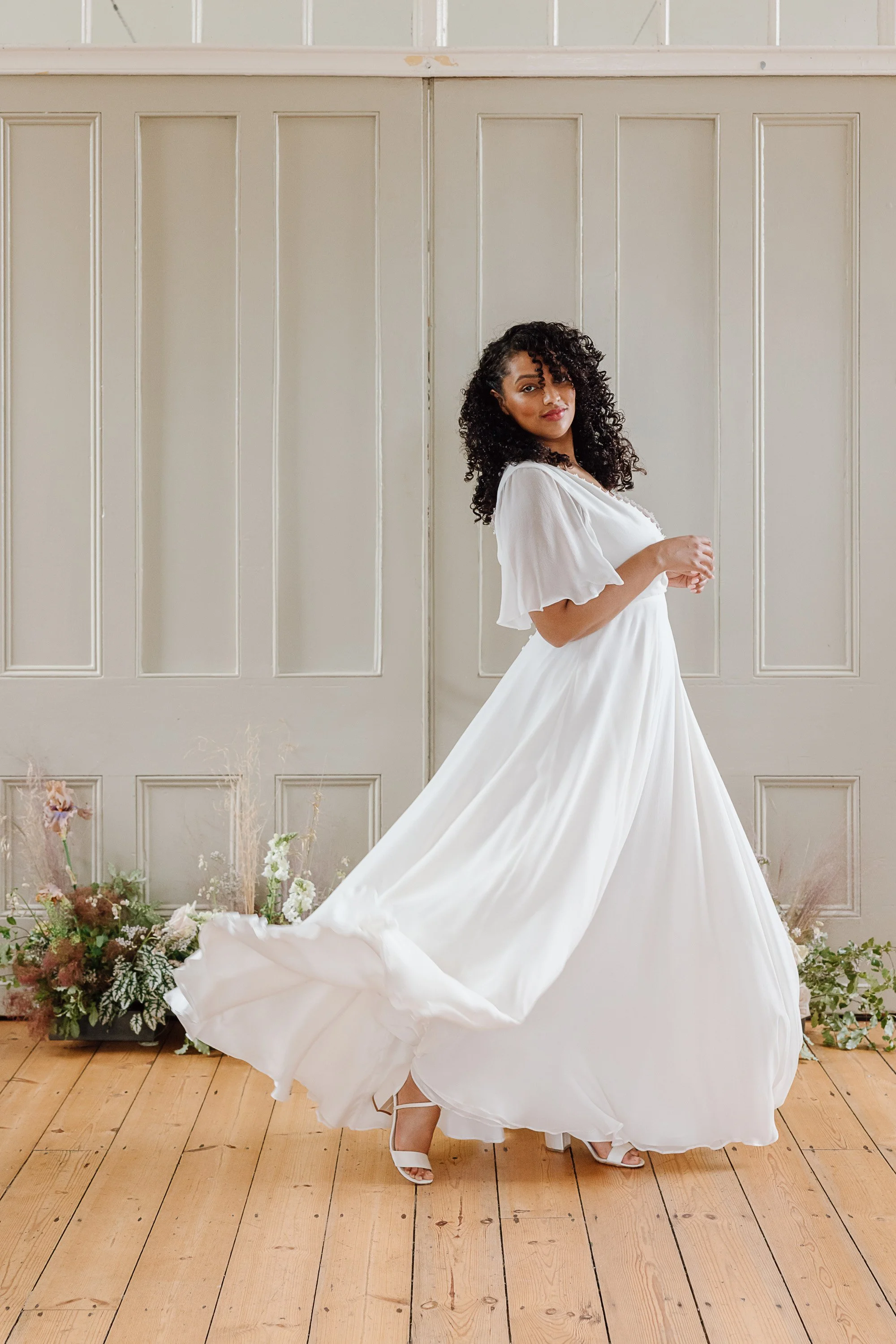 A bride in a flowing white silk gown twirls lightly across a wooden floor, the soft fabric lifting in a graceful arc around her while delicate floral arrangements and tall panelled doors create an airy, romantic backdrop that highlights her poised an