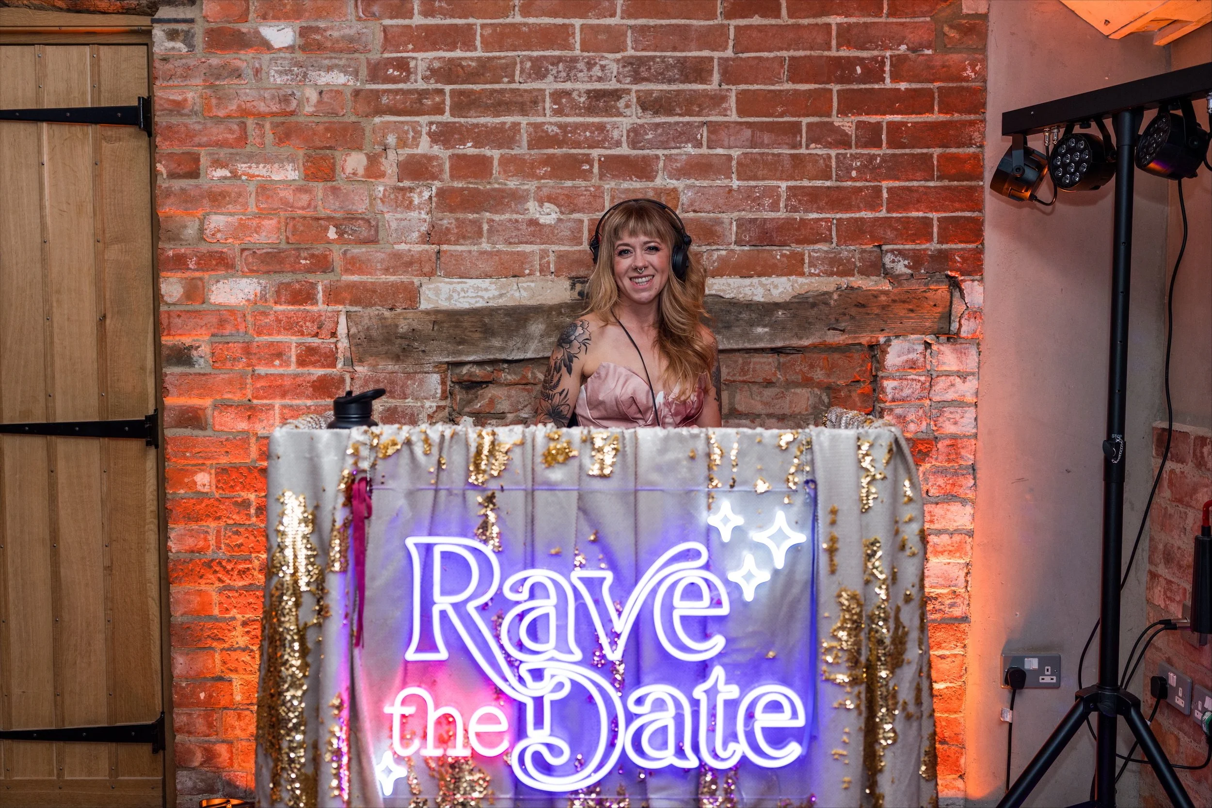 Rave The Date DJ Cara stands at her shimmering DJ booth, framed by warm brick walls and glowing lights. With her headphones on and a bright smile, she radiates energy and confidence behind the signature Rave The Date neon sign. The sparkling gold and