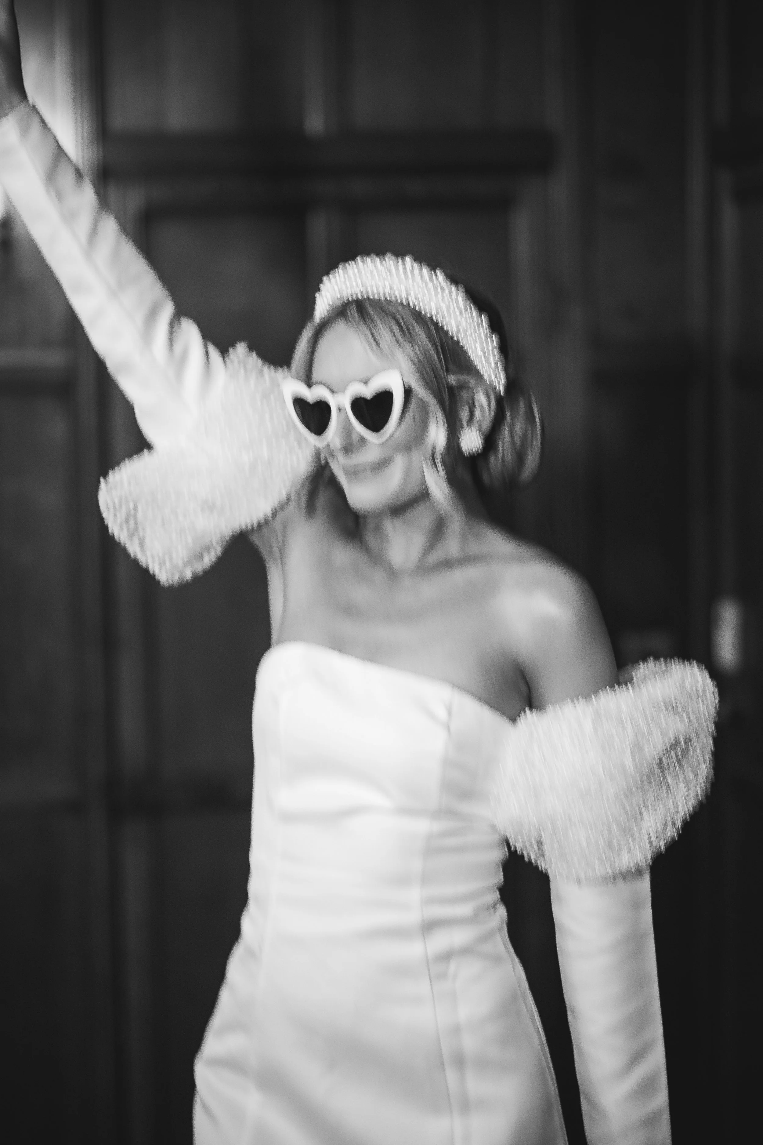 A fun black and white wedding photo captured by Gemma Gaskins Photography featuring a bride dancing in heart shaped sunglasses and a modern off the shoulder gown with textured sleeves. The playful movement, bold accessories and stylish editorial feel