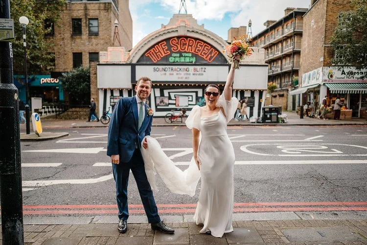 A couple celebrate on a bustling London street outside the iconic Screen on the Green cinema, the bride lifting her bouquet triumphantly while her partner laughs beside her. Her elegant gown and sunglasses pair effortlessly with his sharp blue suit, 