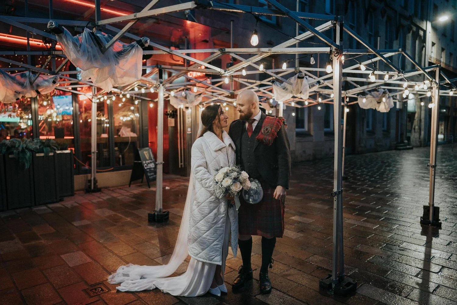A loved-up couple share an intimate moment beneath glowing festoon lights on a rain-slicked Glasgow street, captured in a cinematic, documentary style by Good Luck Wolf wedding photography, blending urban atmosphere, emotion and authentic connection.