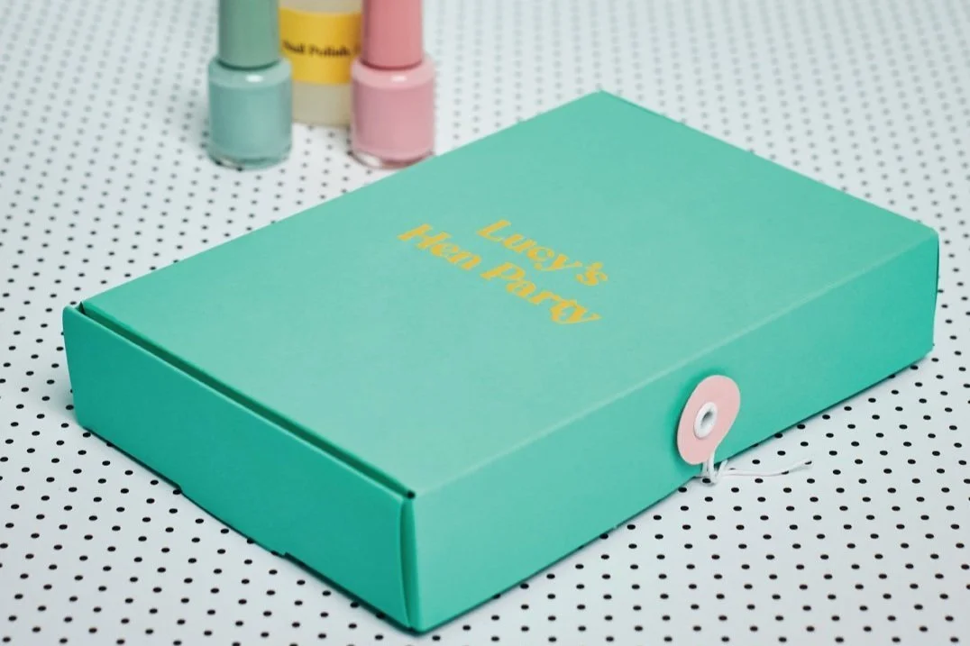 Turquoise gift box with 'Lucy's Hen Party' printed in yellow, designed by Melon Teaspoon for personalized party packaging, with nail polish bottles in the background.