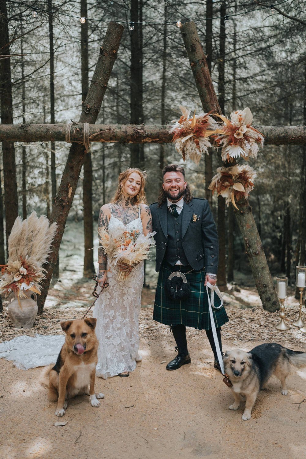 A woodland wedding portrait captured by Good Luck Wolf Photography, showing a tattooed bride in a lace gown and a groom in a traditional Scottish kilt standing beneath a rustic forest ceremony arch, joined by their two dogs for a relaxed, nature-fill