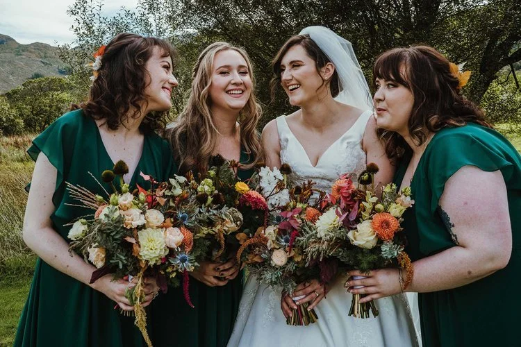 Joyful bridal party holding vibrant autumnal bouquets by New Bud Floristry featuring dahlias, roses, grasses, seed heads and textured seasonal foliage. Set against a scenic countryside backdrop with bridesmaids in deep green dresses, this natural rus