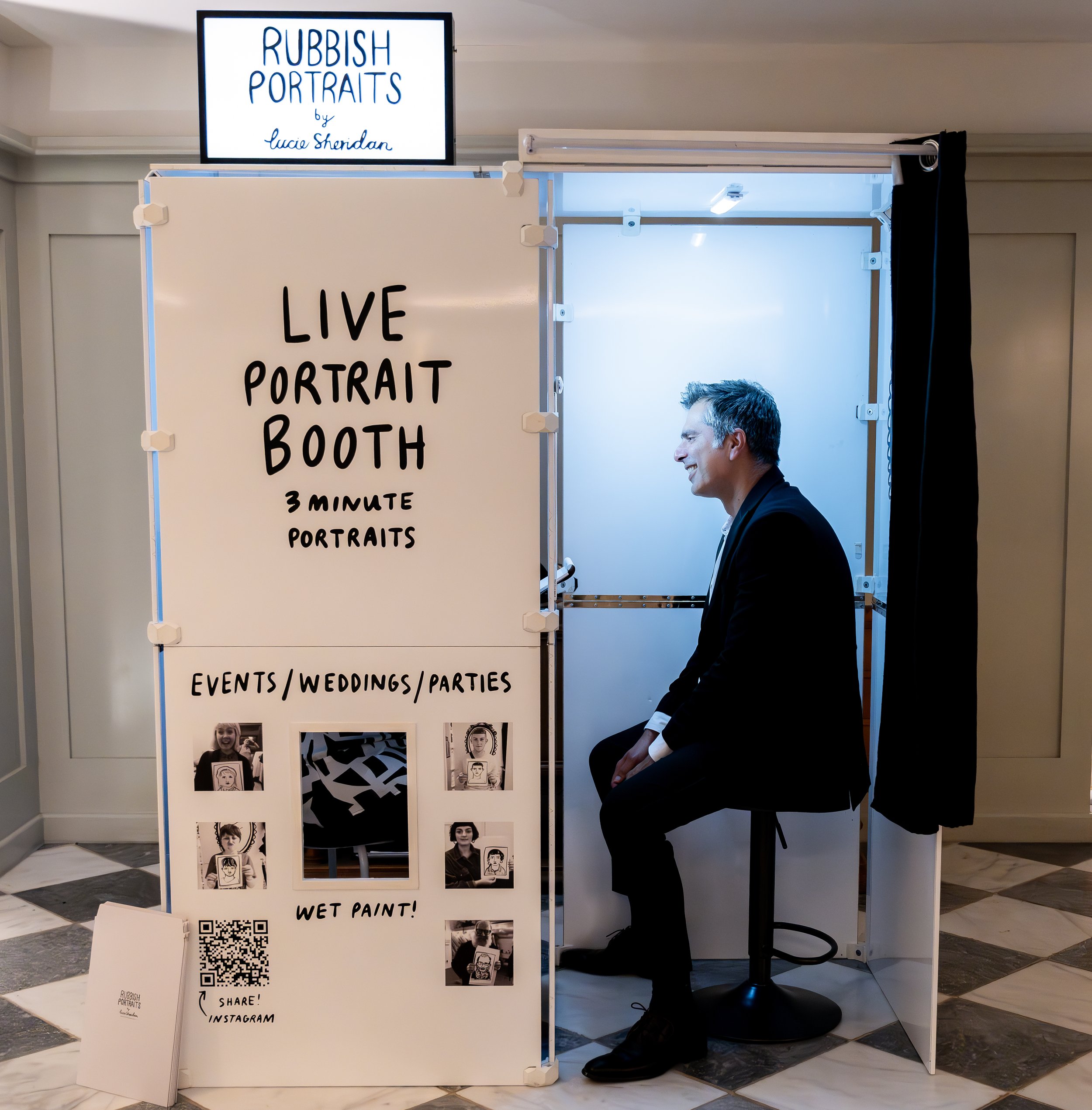 Man sitting inside the ‘Rubbish Portraits’ live illustration booth by Lucie Sheridan, ready for a quick 3-minute portrait session. The booth features a minimalist white design with black lettering and sample artwork, promoting creative entertainment 