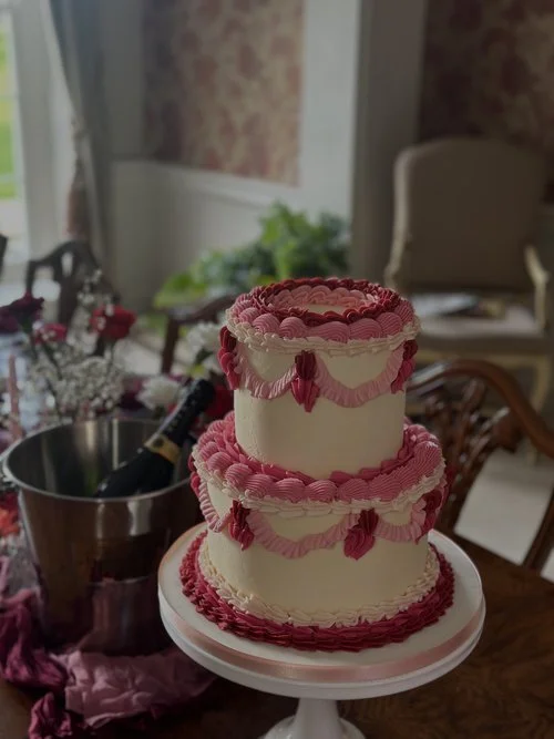A stunning two-tier vintage-style cake by Amber’s Cakes and Treats, featuring bold pink and deep red buttercream piping in elegant swags and ruffles. This statement design channels retro charm with modern precision, showcasing Amber’s signature craft