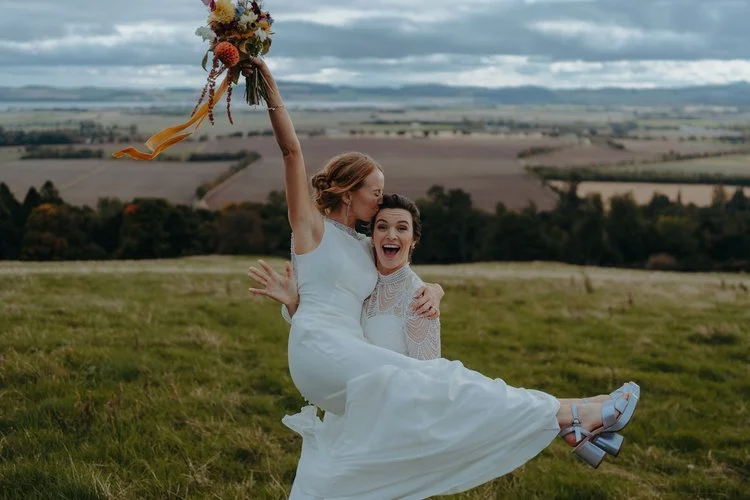 Joy in its purest form, captured with F.D Young Photography’s unmistakable flair. One bride sweeps the other off her feet—quite literally—as they burst into laughter against a sweeping countryside backdrop. The bouquet flies high, dresses flow in the