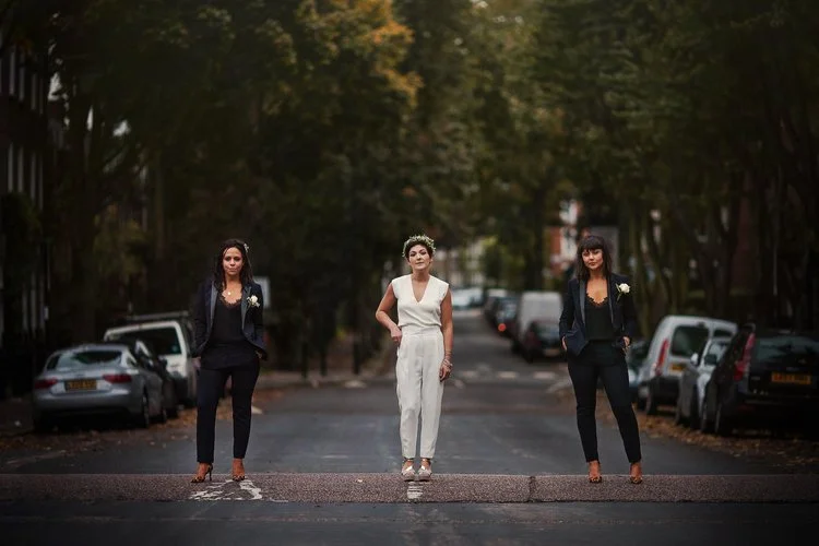 A dramatic portrait set in the middle of a quiet, tree-lined street. Three women stand spaced evenly across the frame. At the center, a woman in a white sleeveless jumpsuit wears a floral crown and white heels, her expression calm and confident. On e