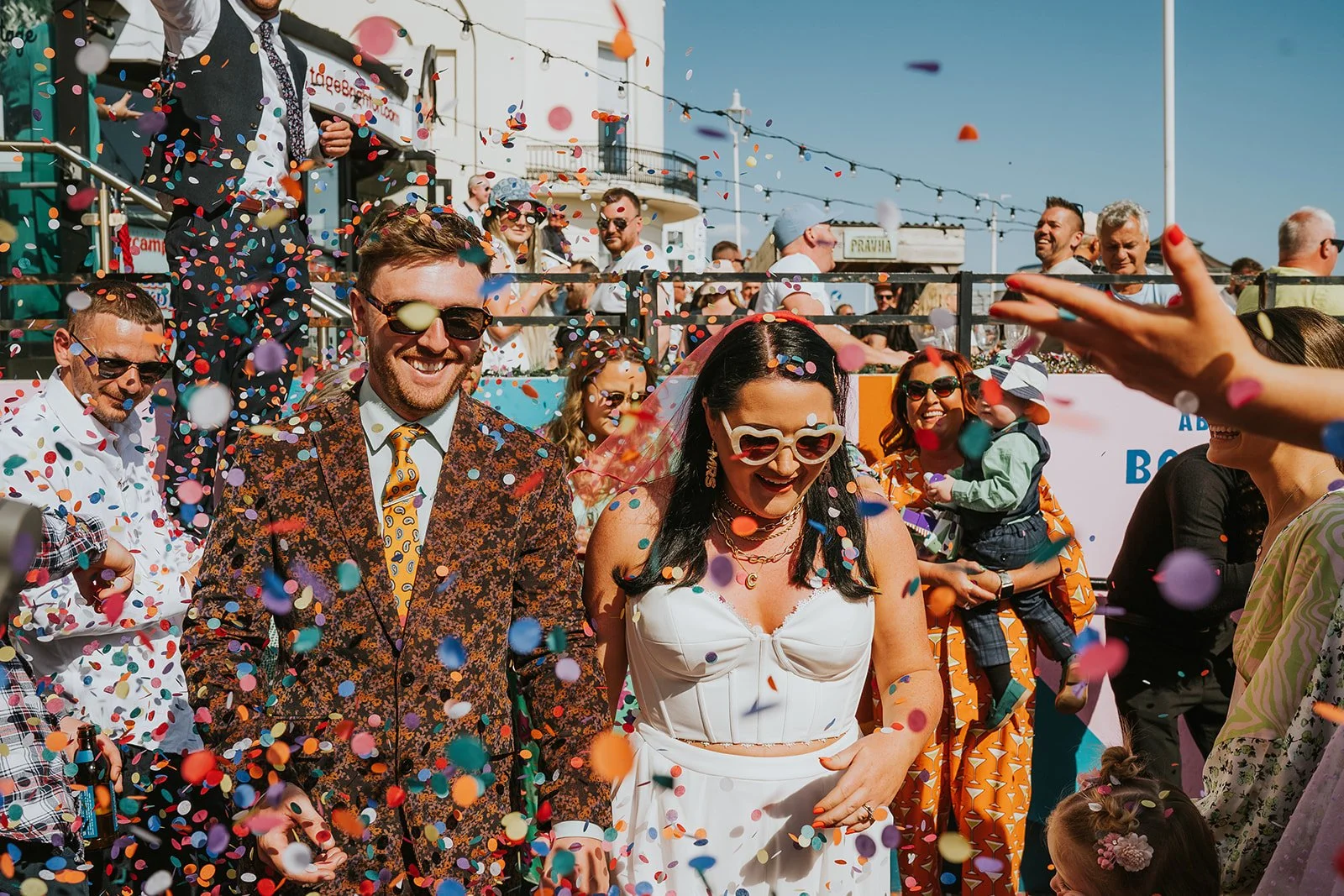 Bride and groom walk through confetti at Pier Court Wedding Venue in Brighton, capturing a joyful, colourful seaside wedding celebration with guests.
