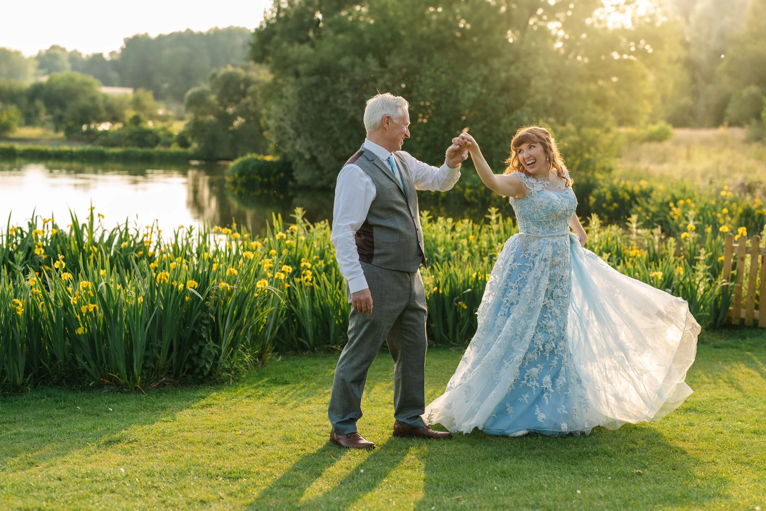 A couple share a joyful dance in the golden evening light beside a serene lakeside garden, beautifully captured by Christy Photography. The bride’s flowing blue lace gown catches the sunlight as they twirl among tall yellow irises, creating a warm an
