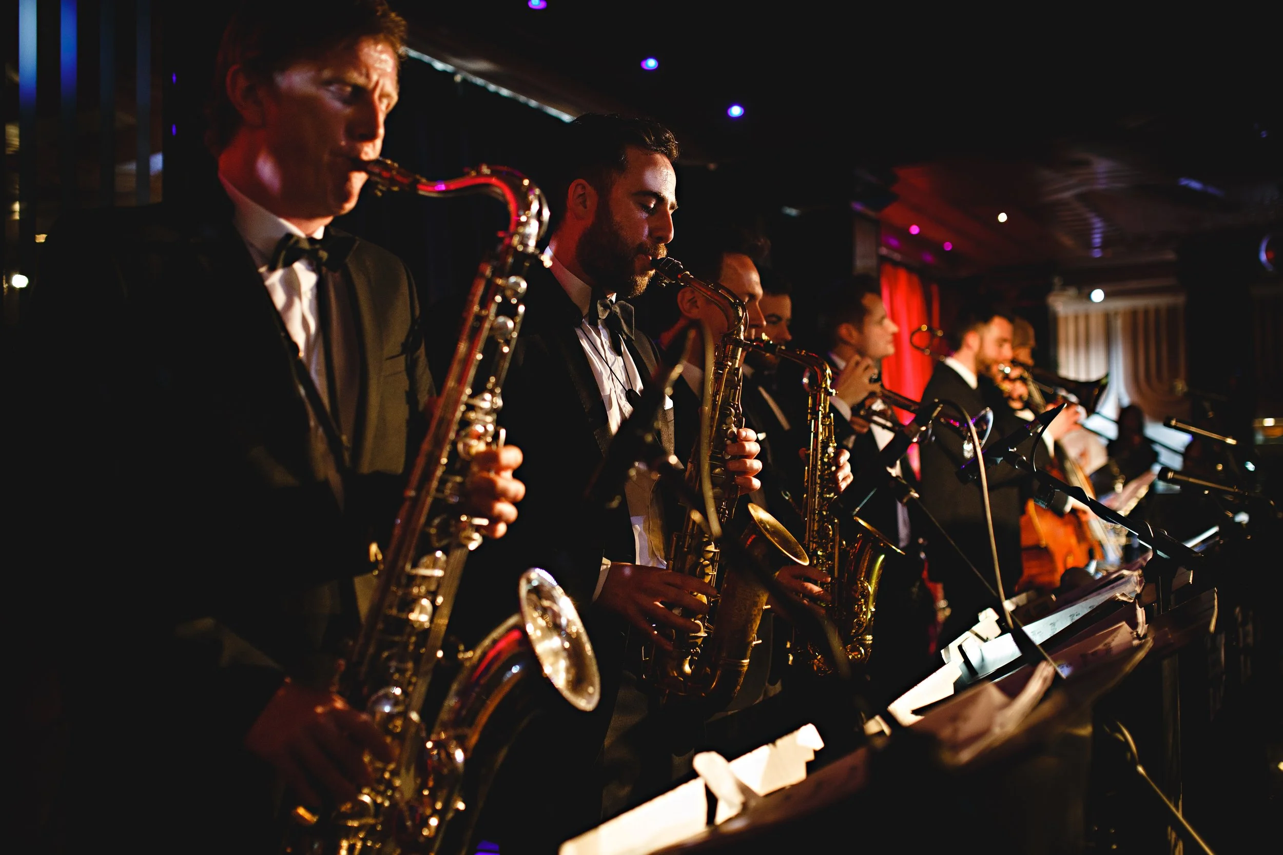 A live horn section from Married to the Music performs at an elegant wedding reception, creating a high-energy atmosphere with smooth jazz, soul, and pop hits. Dressed in black-tie attire, the musicians play saxophones and brass instruments under war