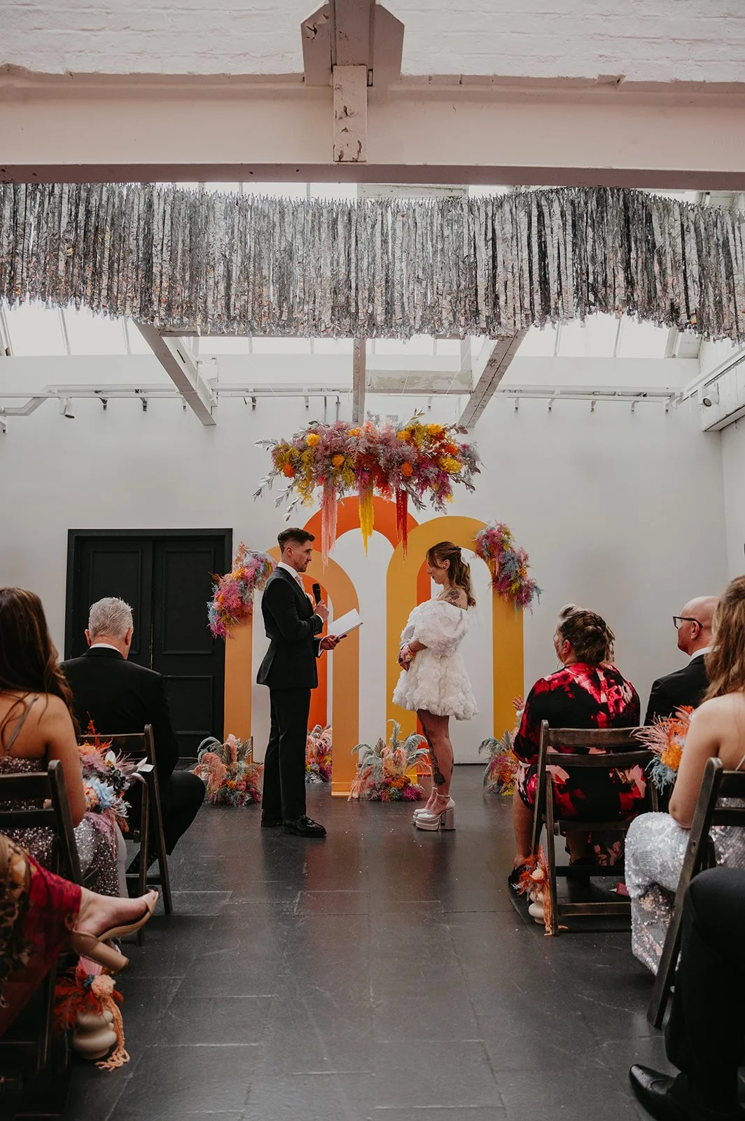 Modern indoor wedding ceremony by AMW Events featuring a bold yellow and orange geometric arch with vibrant floral arrangements and a hanging installation, as the couple exchanges vows under silver tinsel garlands.