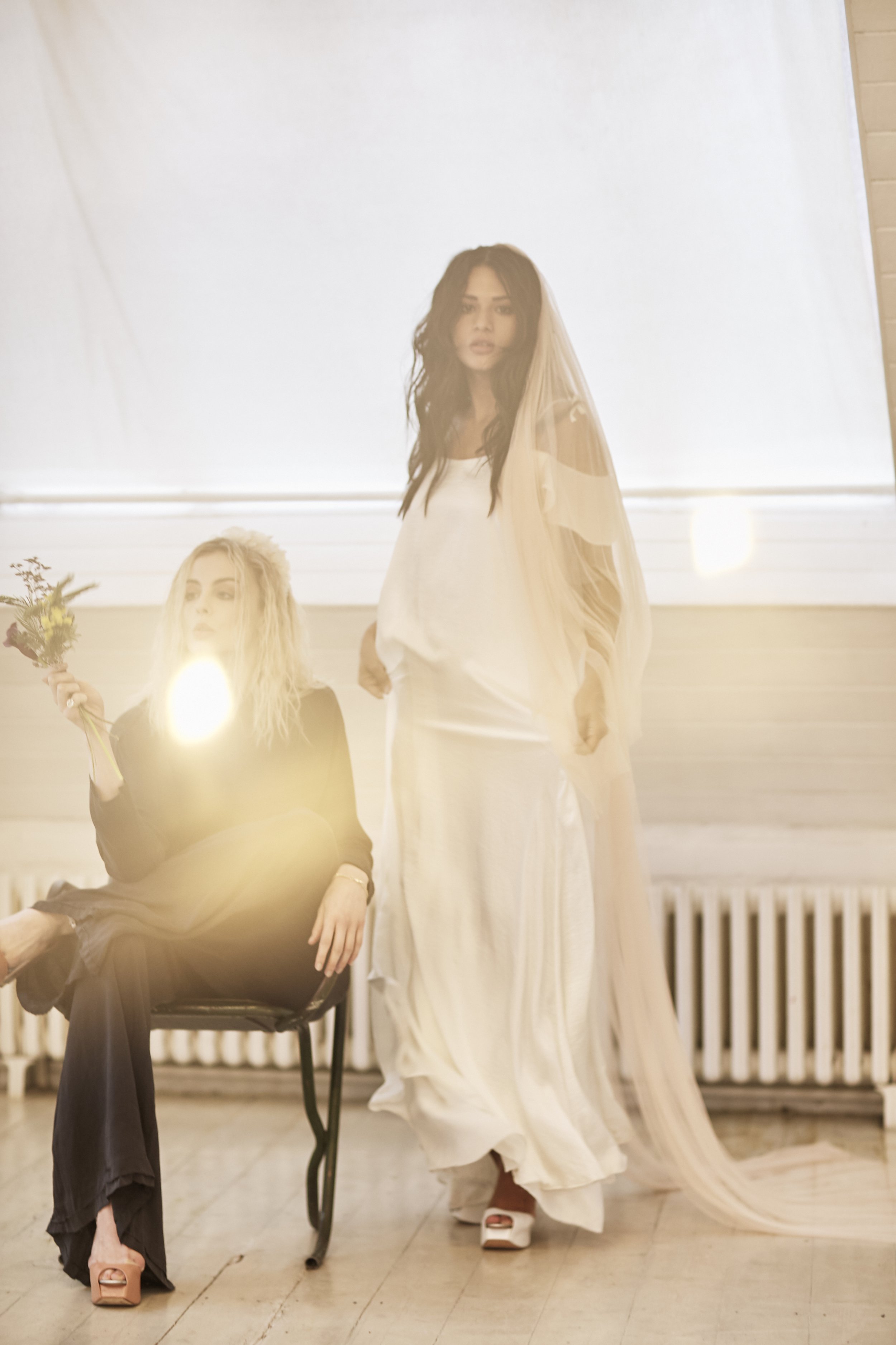 Two figures are arranged in a softly lit interior, one seated and one standing, partially veiled by warm flare from the light. Sheer layers fall loosely over simple silhouettes, with the standing figure’s long, translucent skirt and veil catching the