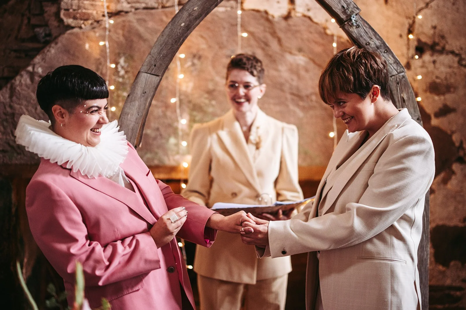 A joyful same-sex wedding ceremony in the UK featuring two brides exchanging rings under a rustic wooden arch adorned with fairy lights. Both wear stylish tailored suits - one in pink and one in cream - while their wedding celebrant Chloe Green offic