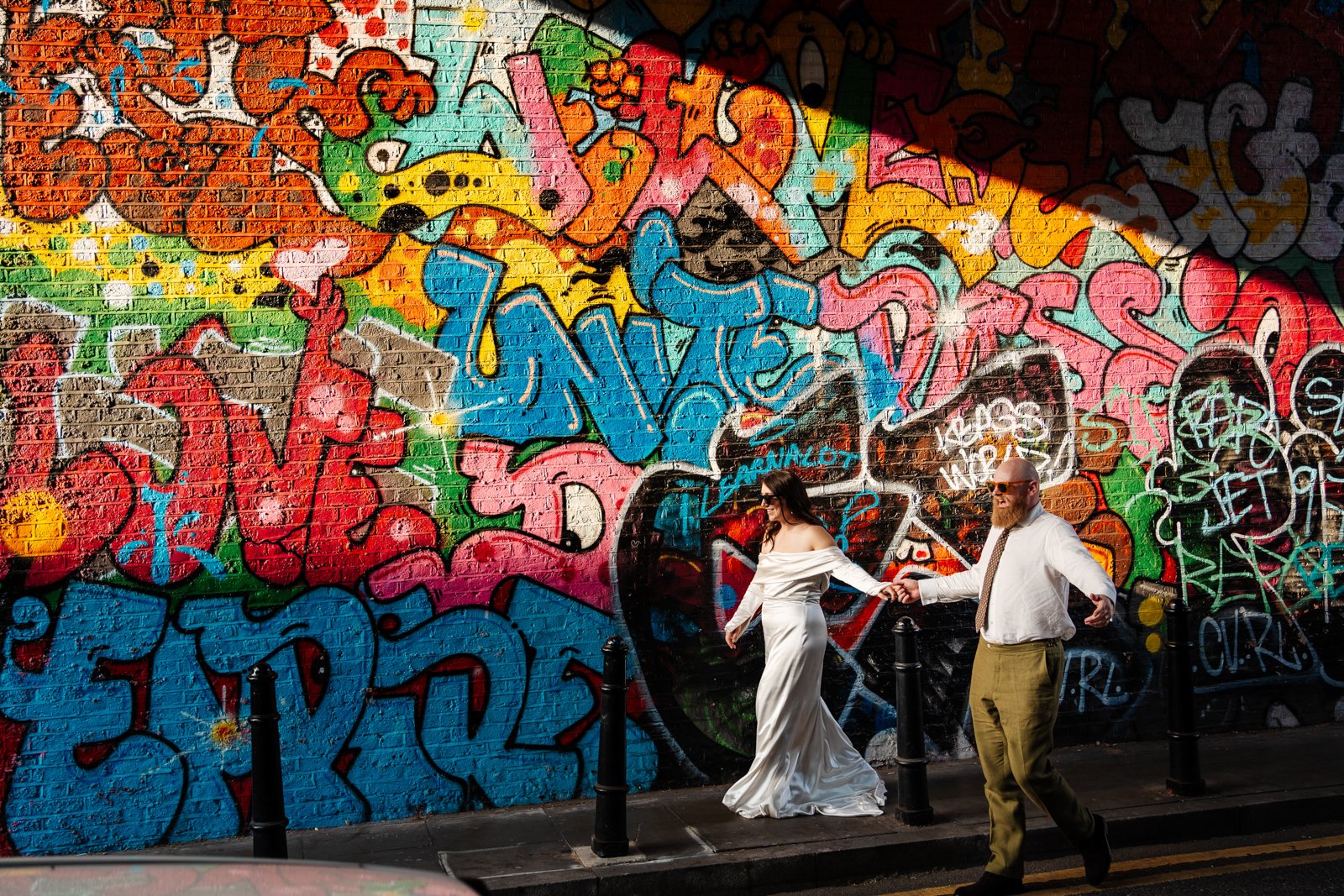 A couple walks hand in hand past a brightly coloured graffiti-covered brick wall, with the woman wearing a long off-the-shoulder white dress and the man dressed in a white shirt and tie with olive trousers. Sunlight creates dramatic patches of light 