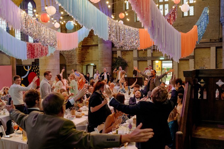 An energetic wedding celebration styled by Streamadelica Event Styling. The grand venue is transformed with hanging pastel fringe garlands, metallic streamers, and colourful paper lanterns suspended from the high ceilings. Guests dance and celebrate 