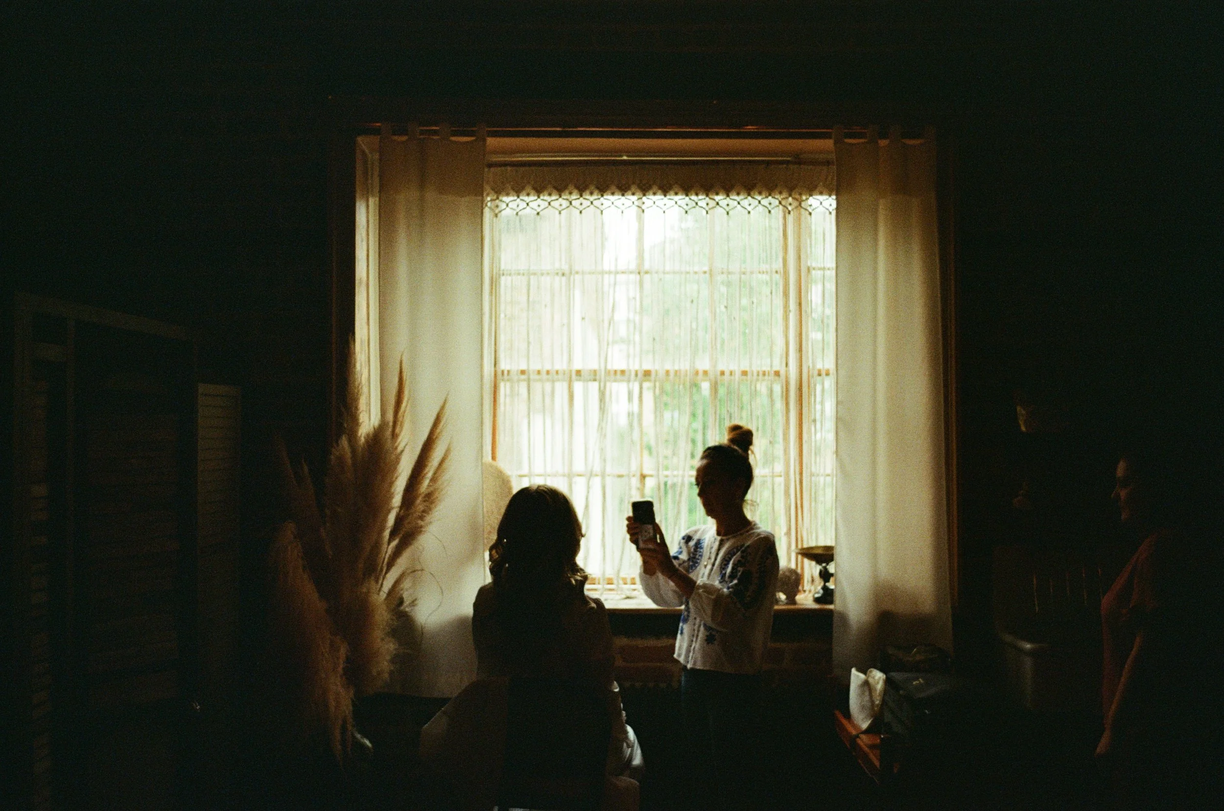 A bride prepares for the day in a softly lit room, captured in the warm grain of 35mm film as natural light pours through delicate lace curtains. Silhouetted against the glowing window, she sits calmly while a friend or family member documents the mo
