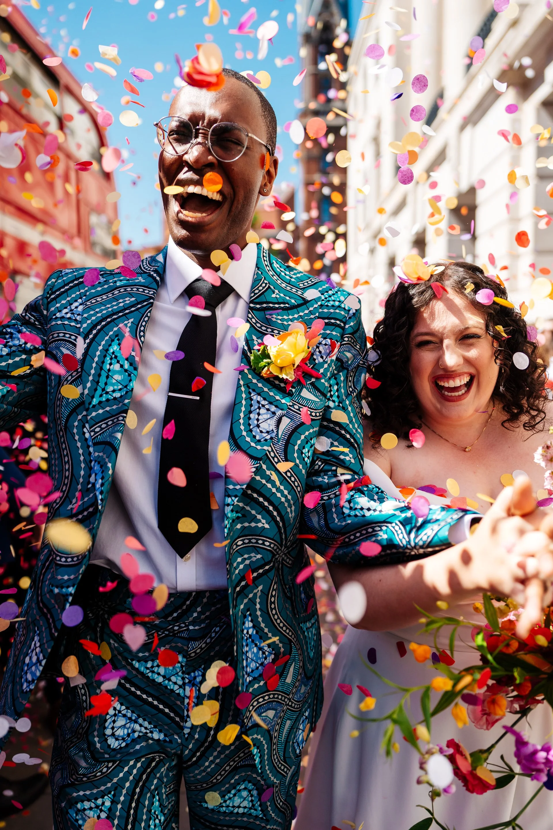 A joyful couple is pictured walking through a busy street as vibrant multicoloured confetti is thrown around them, with both individuals laughing and holding hands. The person on the left wears a patterned teal suit with a boutonniere and the person 