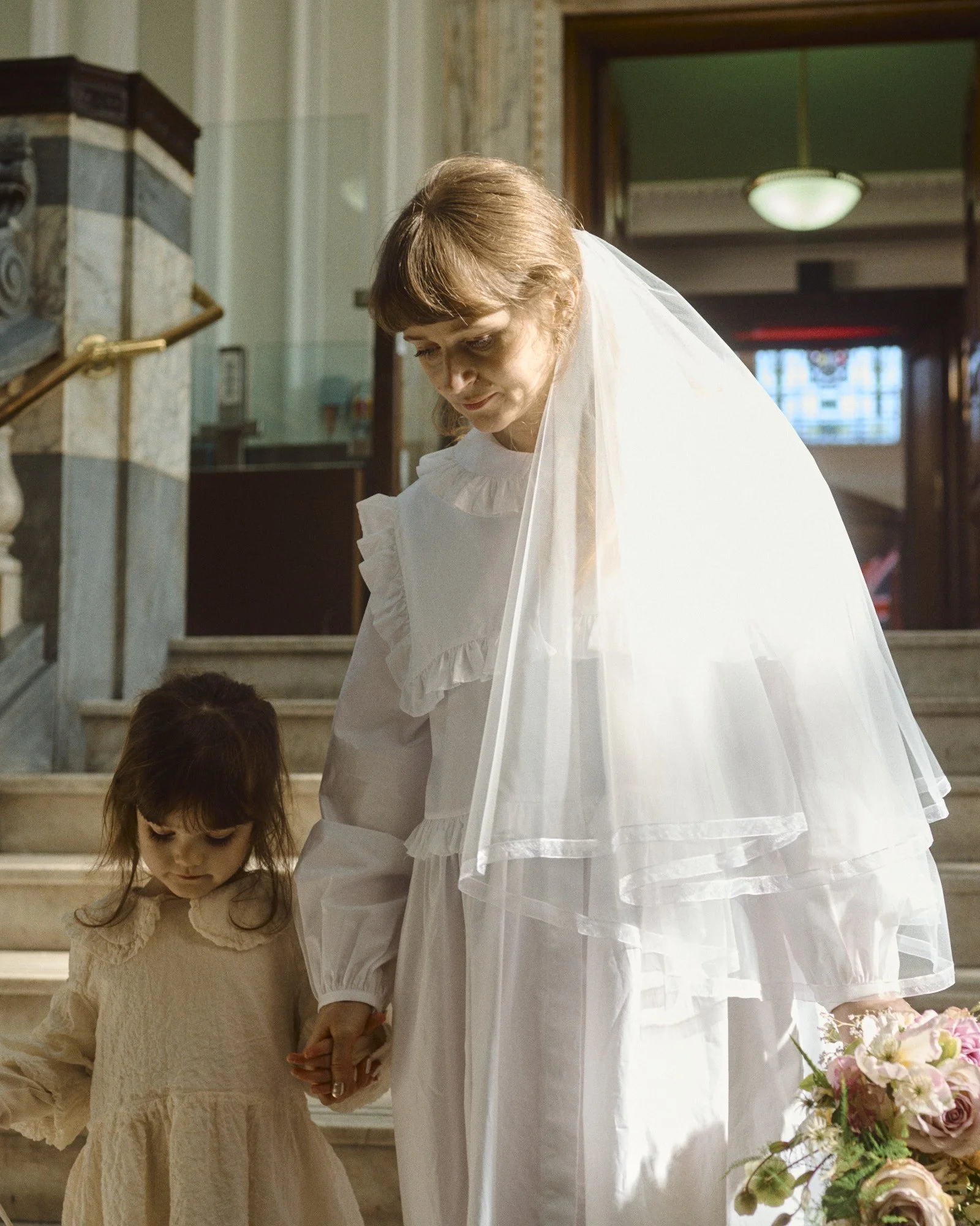 A warm and intimate moment captured by Wayne La Photography, showing the bride and a young flower girl walking hand in hand down a sunlit staircase inside a grand historic building. The bride’s soft, vintage inspired gown and sheer layered veil catch
