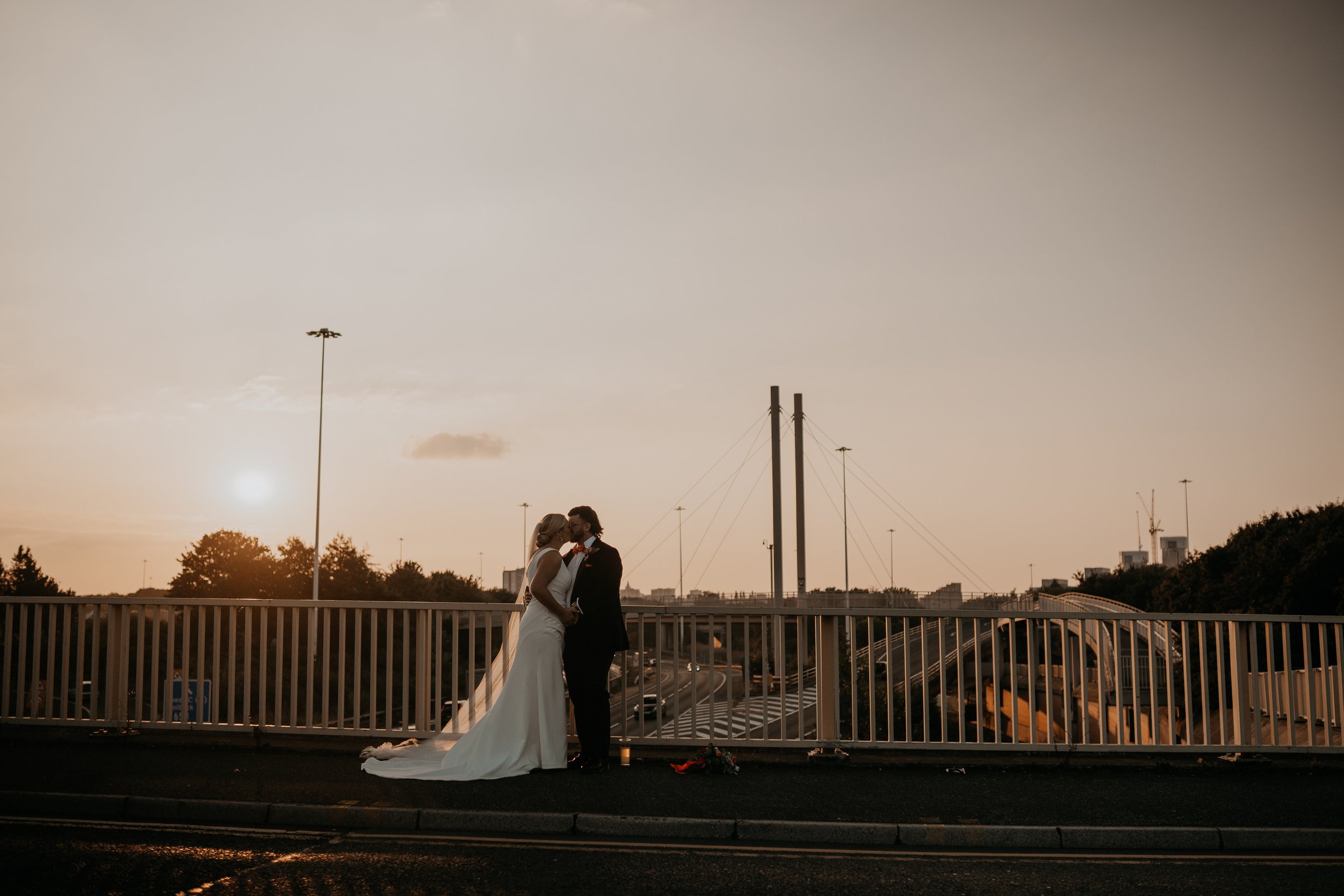 A couple stands on an overpass at sunset sharing a quiet kiss as soft golden light washes across the sky and silhouettes the tall bridge cables and distant city structures behind them, with the bride’s long veil drifting gently toward the ground and 