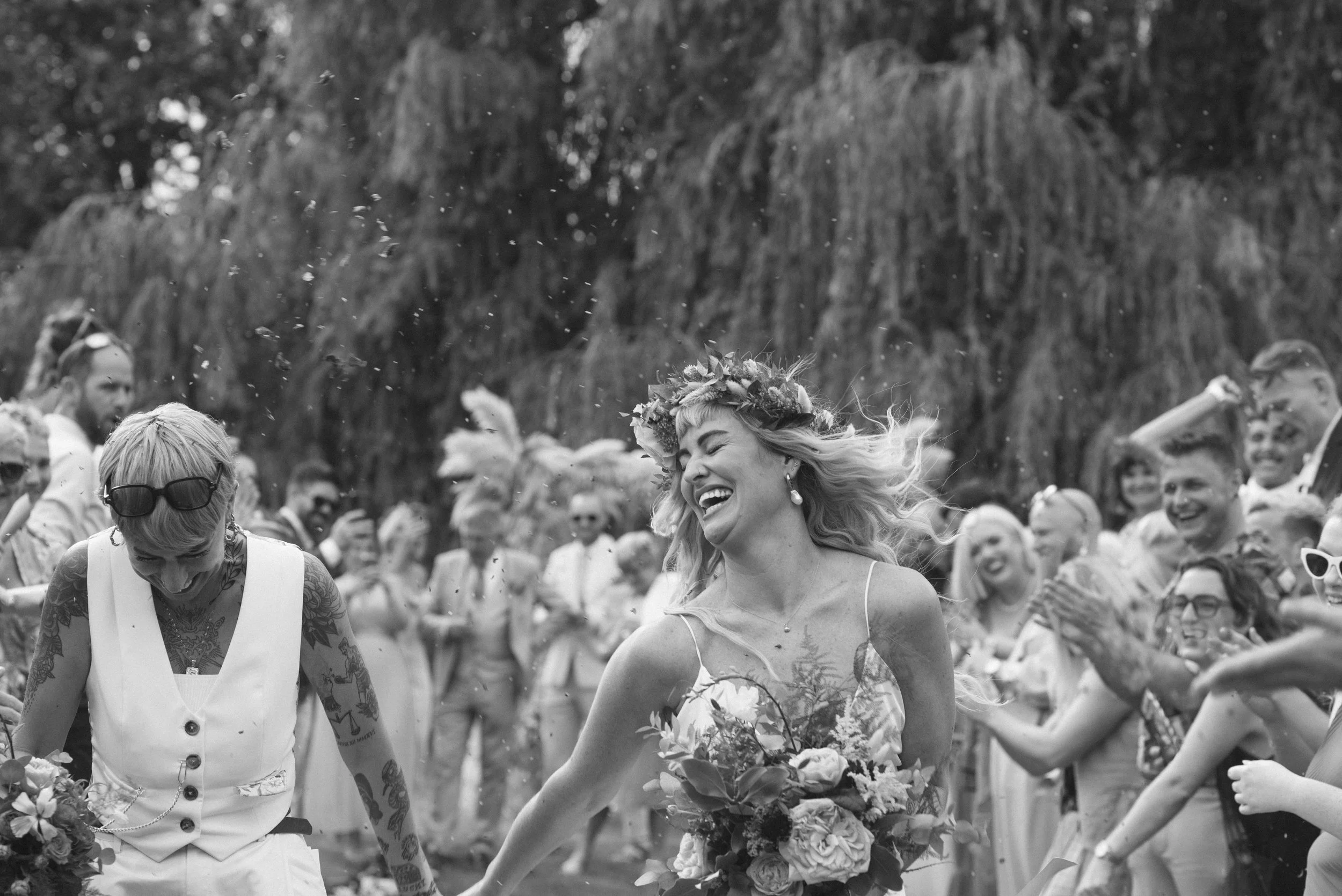 A joyful couple walk hand in hand through a shower of confetti surrounded by cheering guests, captured by Roz Pike Photography. The black and white documentary style highlights their laughter, floral details and the lively celebration atmosphere of a