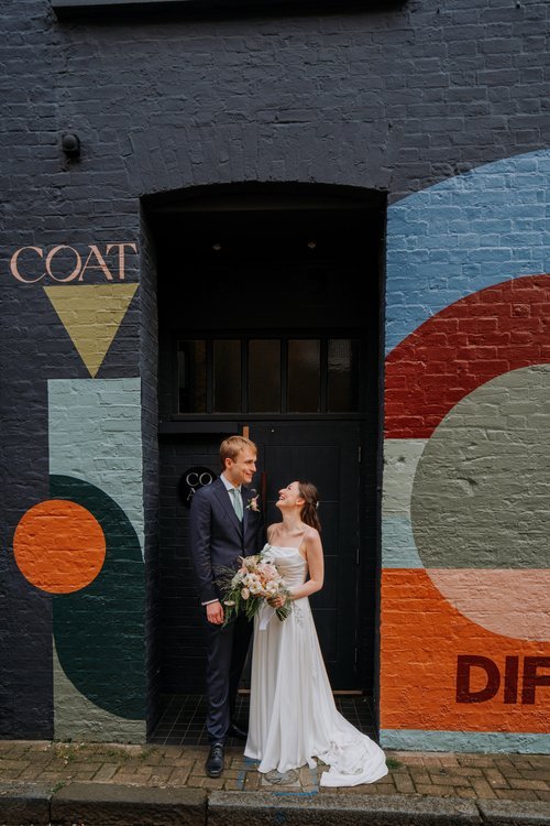 A newly married couple stand smiling at each other in front of a colourful geometric mural in an urban London street, captured by Joshua Humphrey Photography. The bride holds a soft, natural bouquet while the groom looks at her affectionately, creati