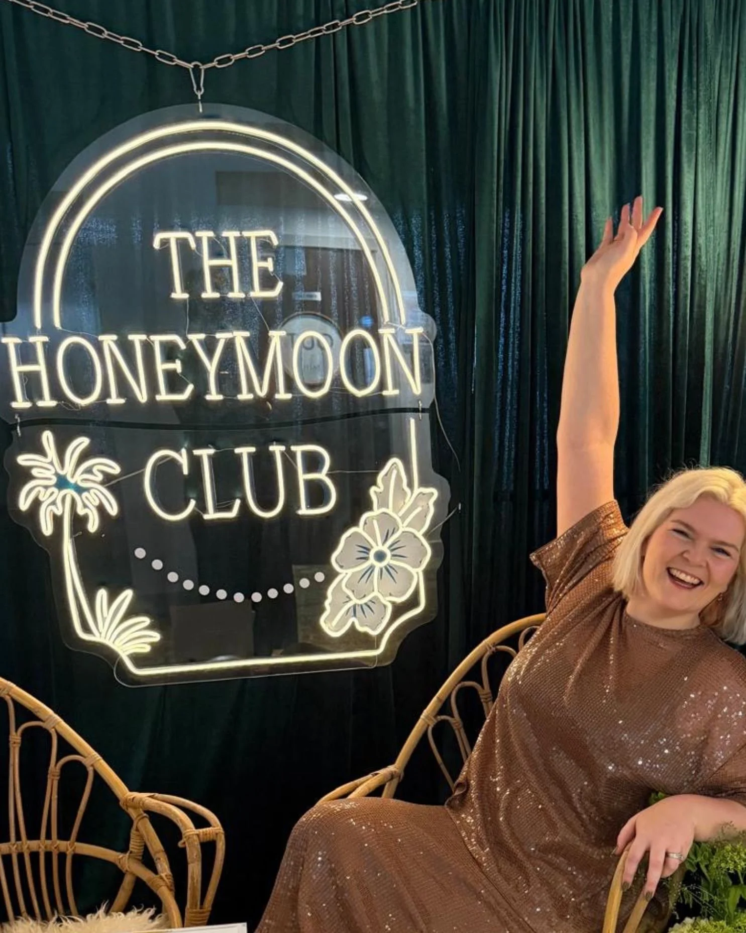 A blonde woman sits in front of a neon sign that says The Honeymoon Club. Her name is Sophie Linton and she is the founder.