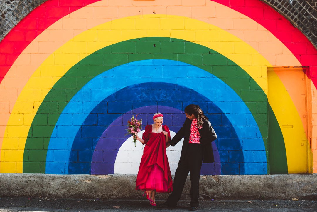A vibrant and modern wedding portrait photographed by Barbara K Photography, featuring a stylish couple standing in front of a bold rainbow mural. The bride wears a striking red dress and holds a bright bouquet, while the groom complements the scene 