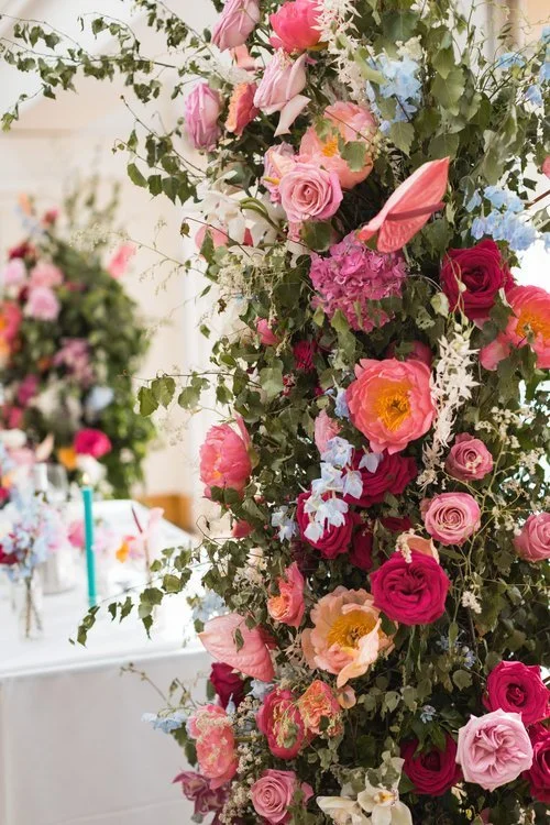 Vibrant floral installation by Studio Clarisa Floristry featuring lush pink, red, and peach roses, anthuriums, and pastel blue blooms with cascading greenery, creating a romantic wedding backdrop indoors.