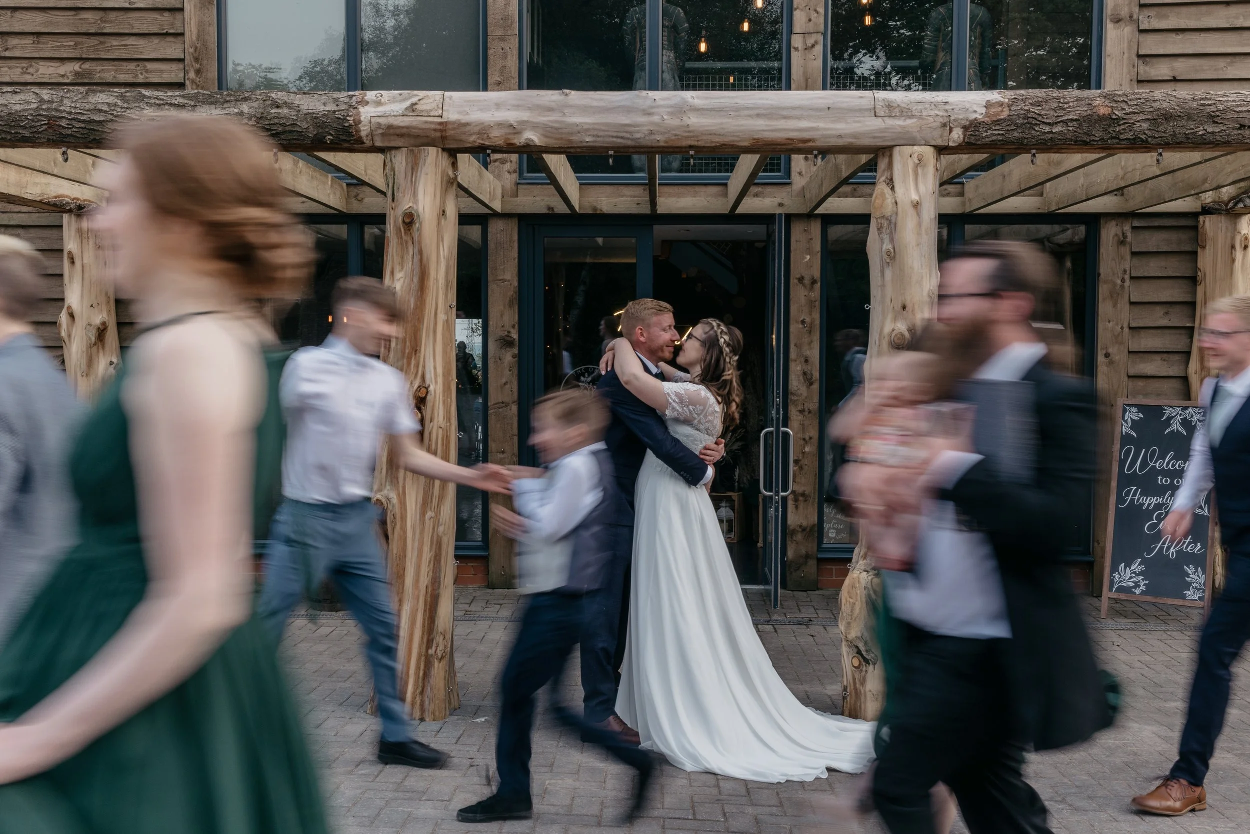 A beautiful, storytelling moment captured by YU Photography, showing the newly married couple sharing a kiss beneath a rustic wooden entrance while the world moves around them in a blur. Guests rush past in soft streaks of motion, creating a dynamic 