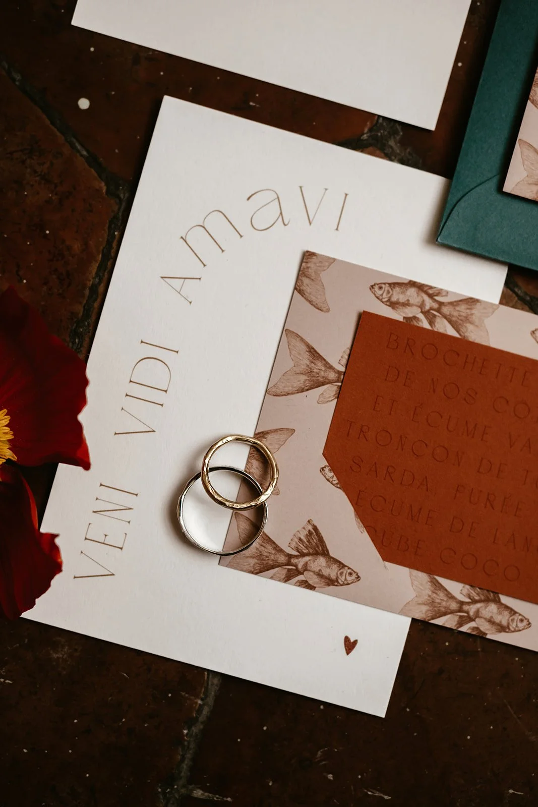 A romantic wedding flatlay featuring Latin inspired stationery reading Veni Vidi Amavi paired with illustrated menu cards in warm earthy tones, rich green envelopes and a pair of textured gold and silver wedding rings styled on rustic terracotta tile