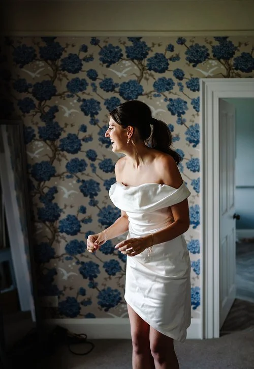 A bride stands in a beautifully patterned room as she gets ready, laughing and full of anticipation while adjusting the off the shoulder neckline of her chic, modern mini dress. Soft natural light highlights the elegant folds of her gown and the warm