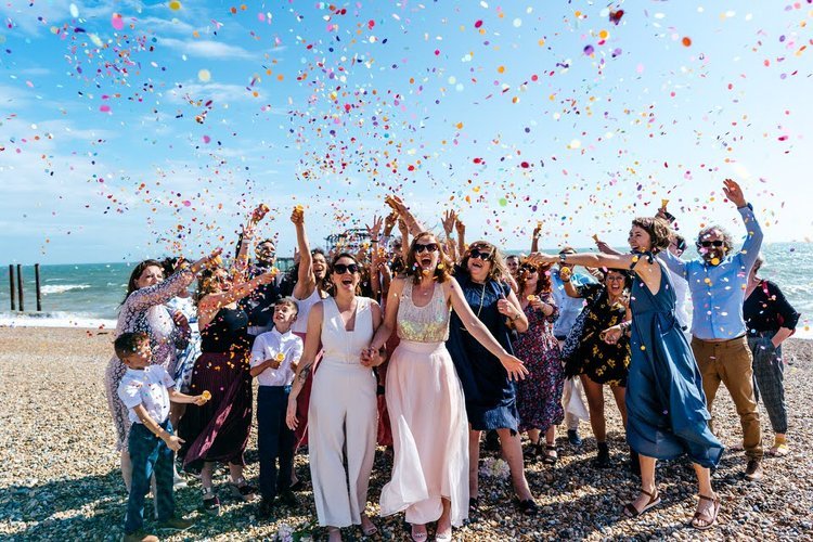 A newly married couple celebrate on a sunny pebble beach as friends and family throw colourful confetti into the air, captured with energy and joy by Jordanna Marston Photography. The bright blue sky, vibrant colours and lively reactions create a dyn
