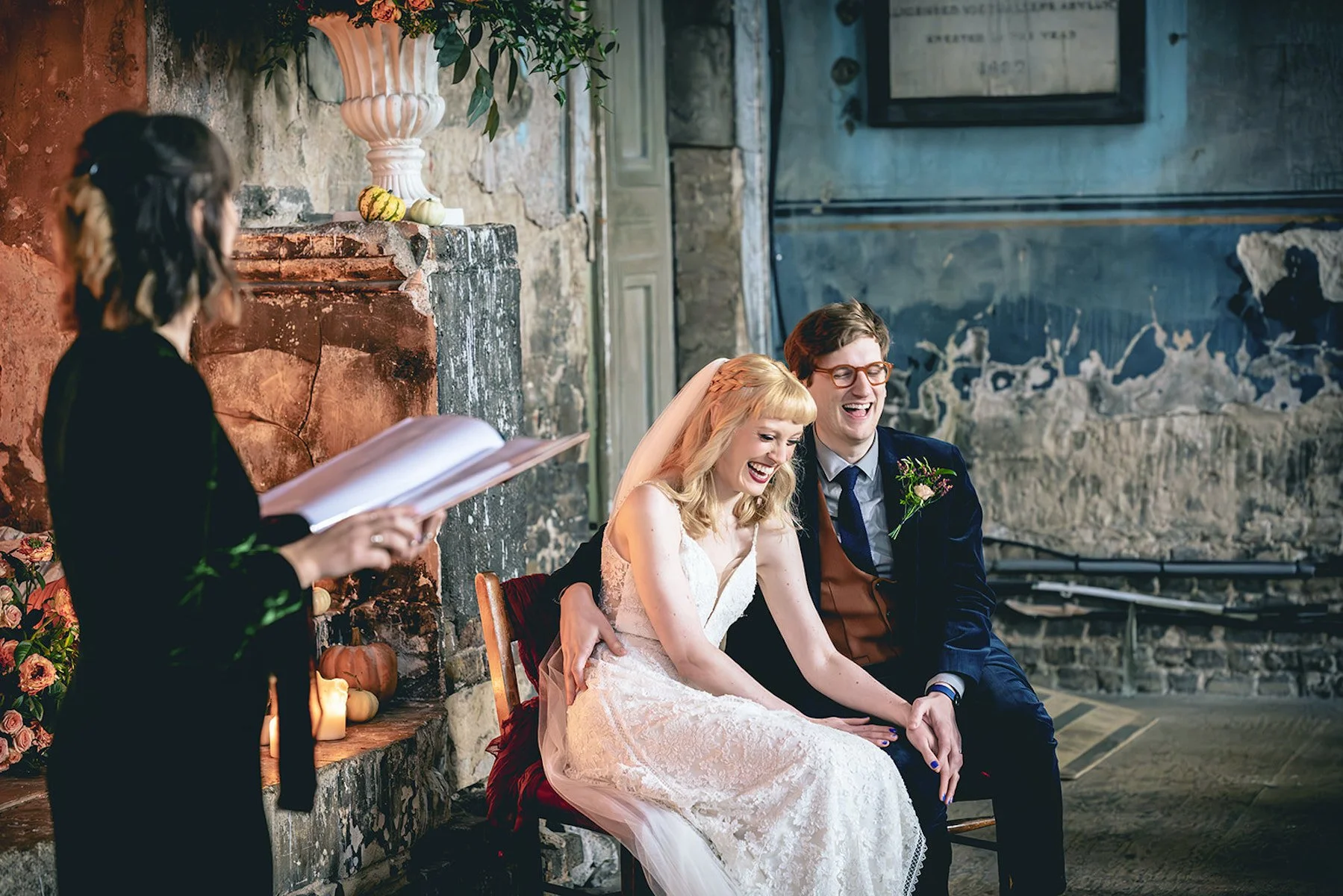 Cherry and Fern Celebrations leads a heartfelt ceremony in a rustic, character-filled venue as the newlyweds share a joyful laugh. The bride in a lace gown and the groom in a navy suit with a rust waistcoat sit together by a vintage fireplace surroun
