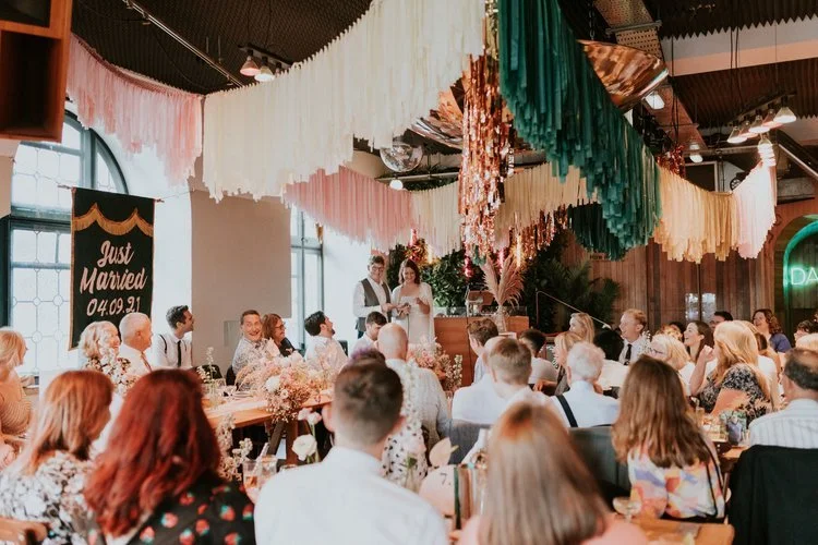 A wedding reception styled by Streamadelica Event Styling. The modern space features cascading fringe garlands in green, pink, and cream, paired with disco balls and soft, romantic lighting. Guests enjoy the lively atmosphere while the couple deliver